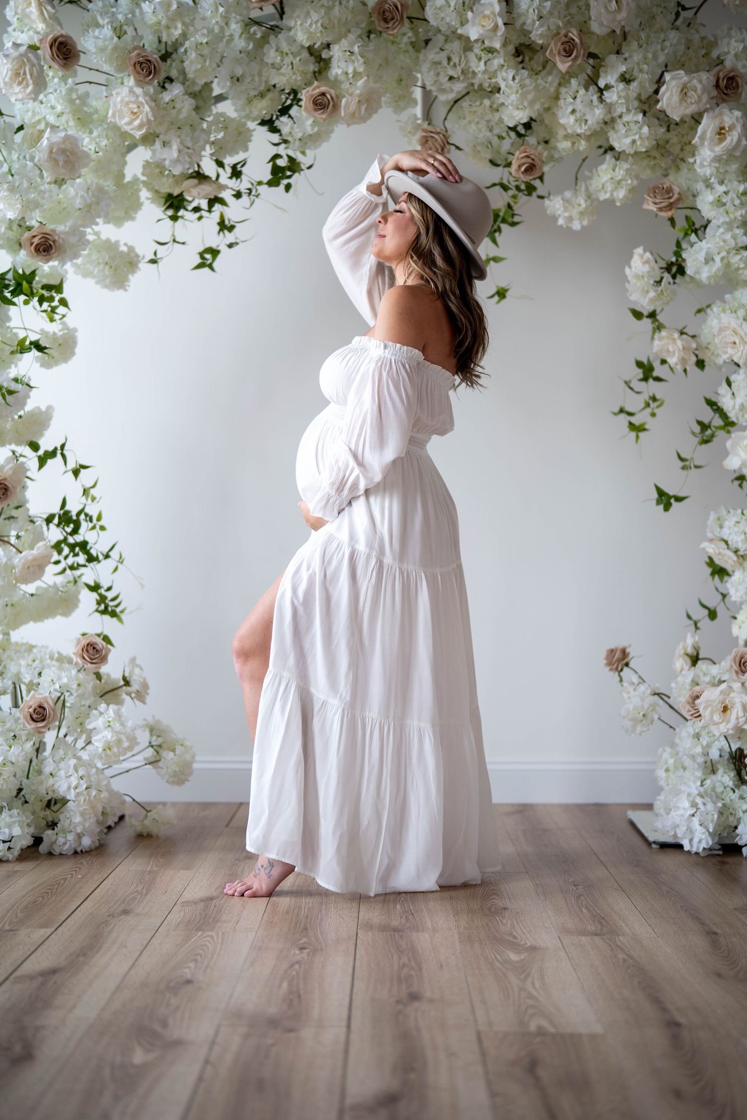 Ethereal maternity pose in white tiered dress against floral backdrop.
