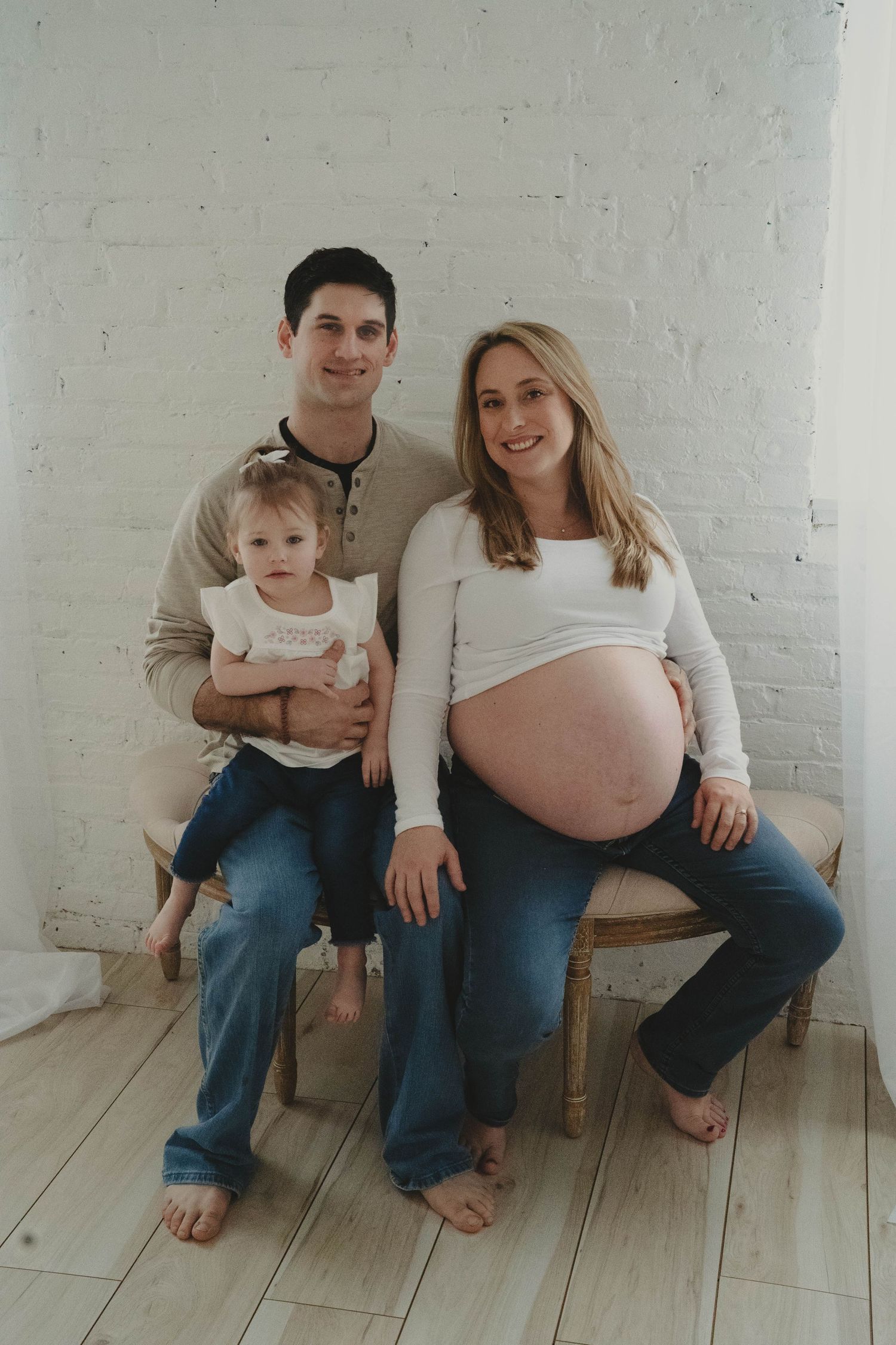Family maternity photos on wooden floor against white brick wall.