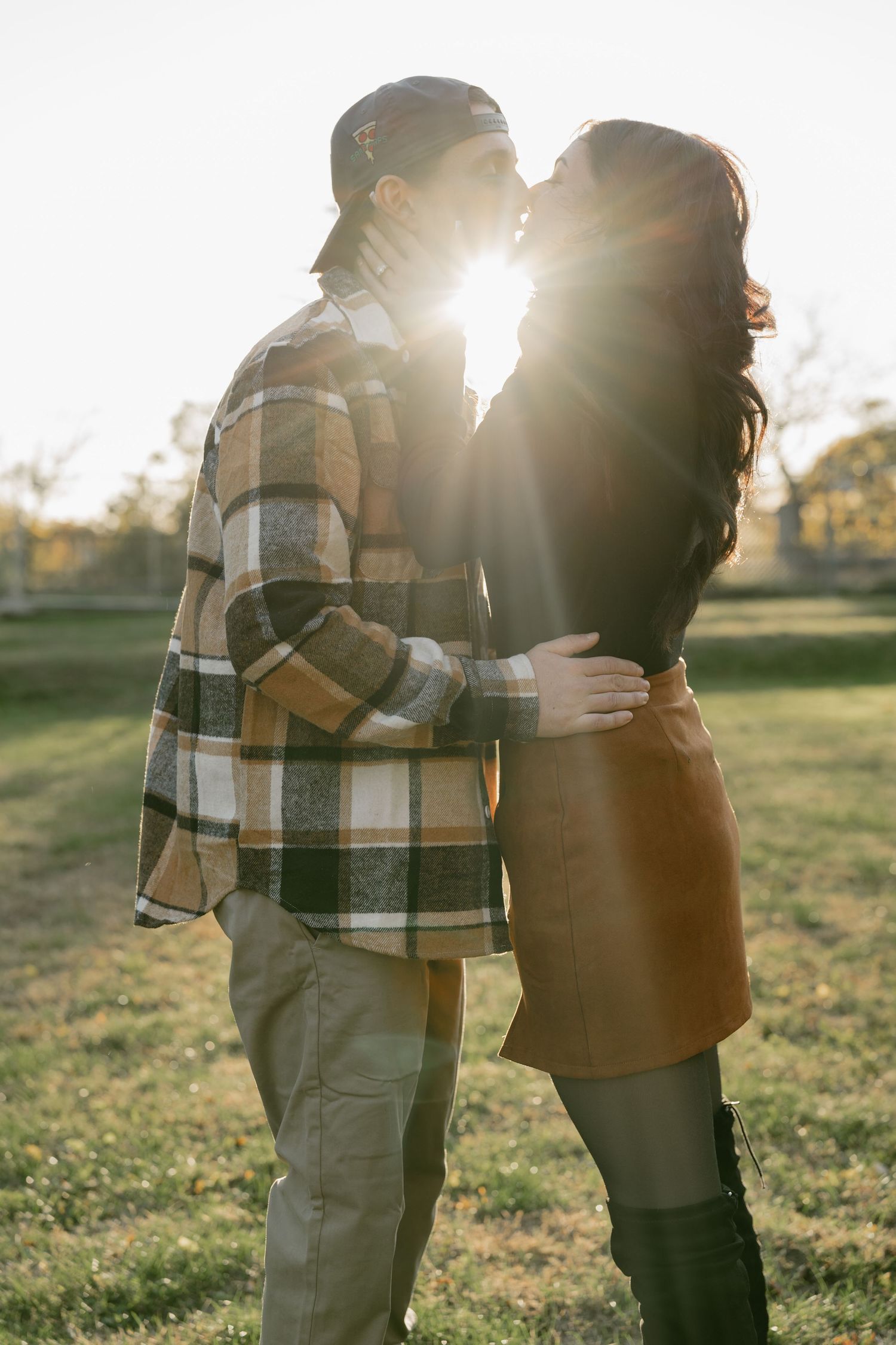 Couple embraces in a sunlit field wearing plaid and casual attire during golden hour.