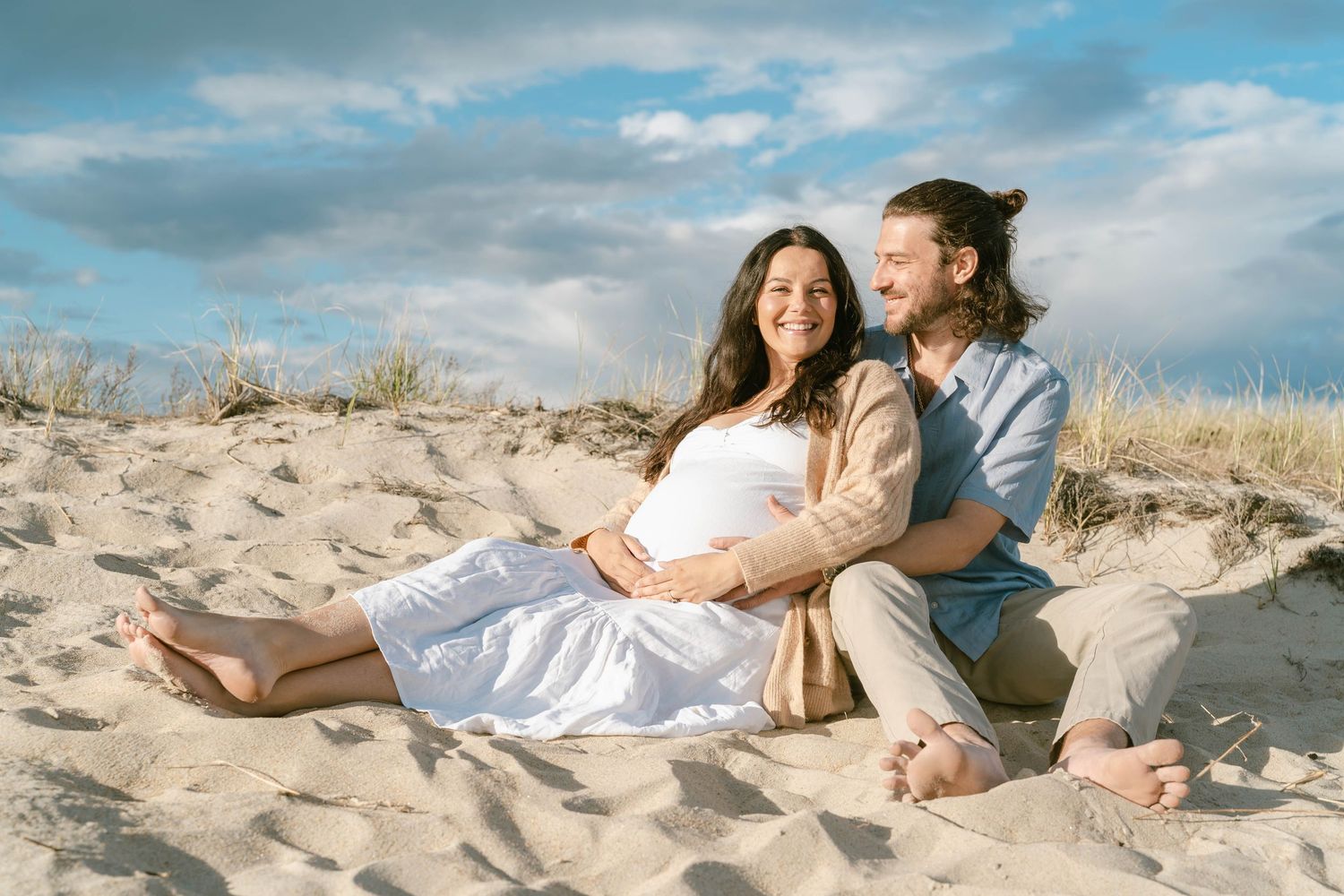 Couple relaxing on beach blanket under blue sky with coastal vegetation.