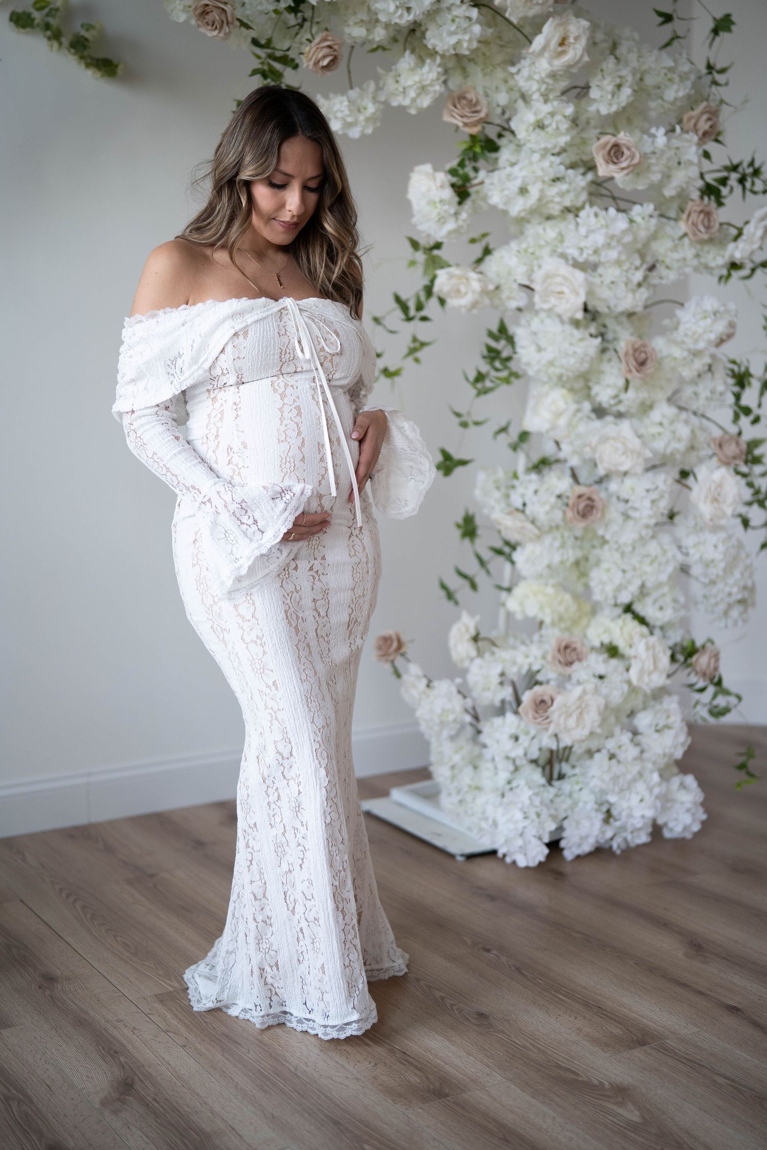 Elegant maternity portrait in white lace gown against floral wall installation.