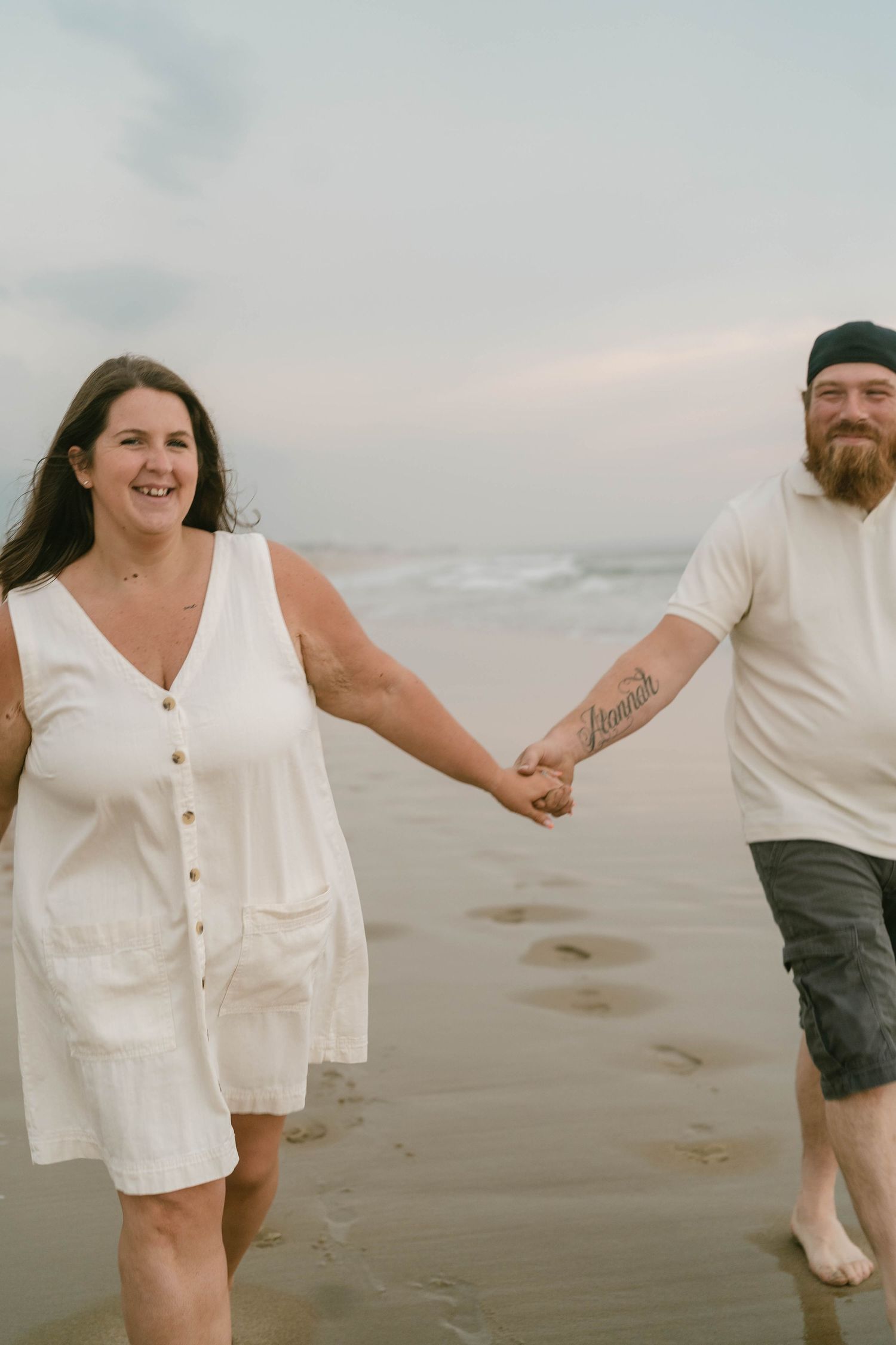 Couple walking hand-in-hand along a sandy beach at sunset, laughing and enjoying a romantic, candid photography session with gentle waves