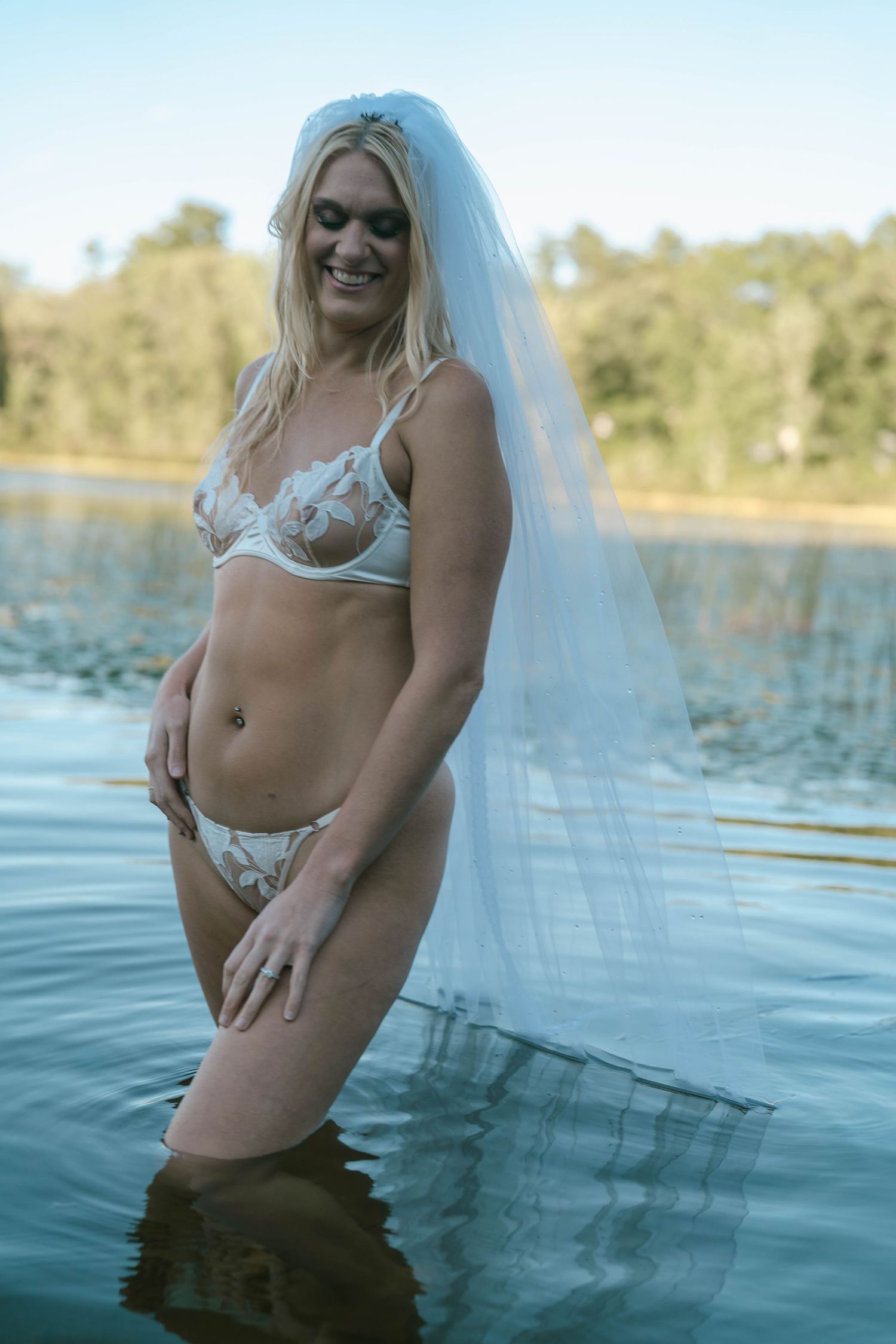 Woman in white lingerie and veil poses in shallow water during outdoor boudoir photoshoot.