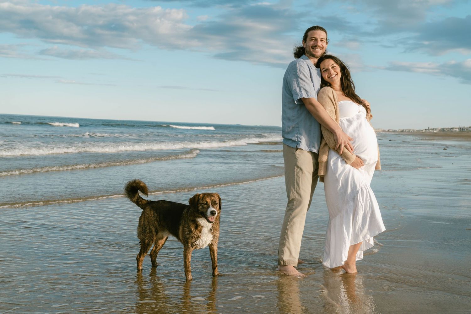 A couple strolls on the beach with their dog during a maternity photoshoot.