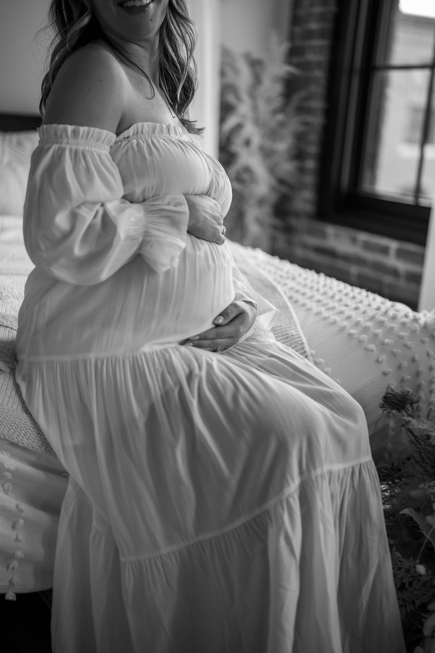 Black and white intimate maternity photo in flowing white dress by window.