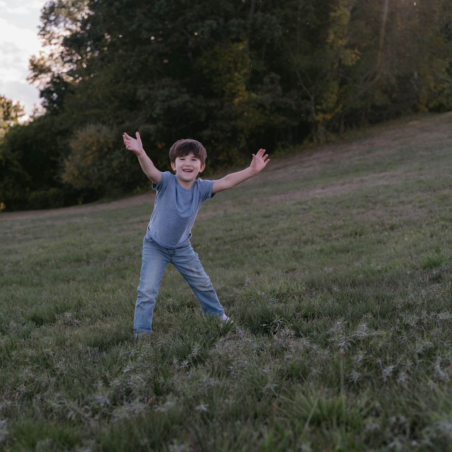 A joyful figure plays on a grassy hillside at sunset with arms outstretched in a playful pose.