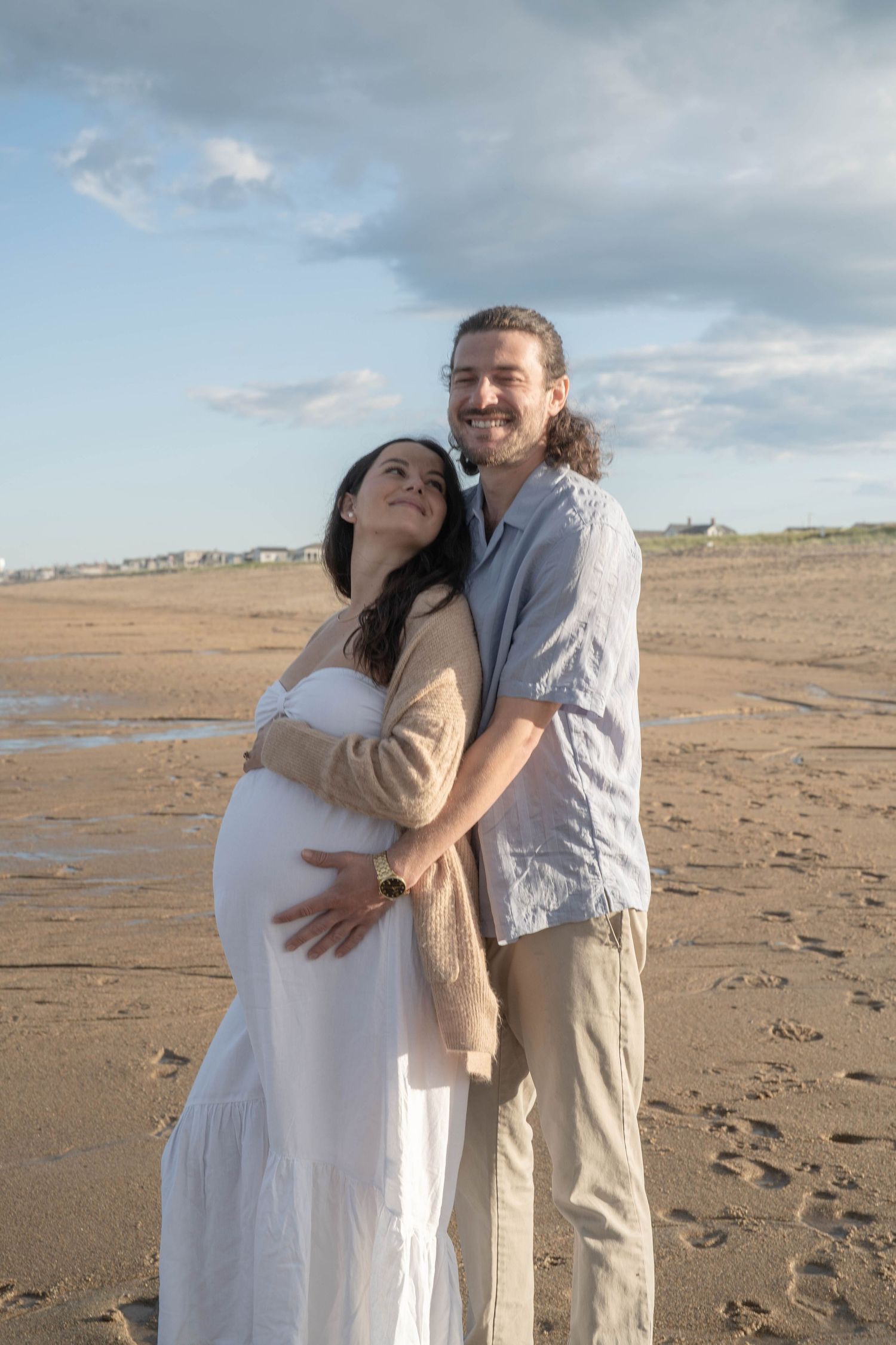 A couple shares a tender moment on an overcast beach during a maternity photoshoot.