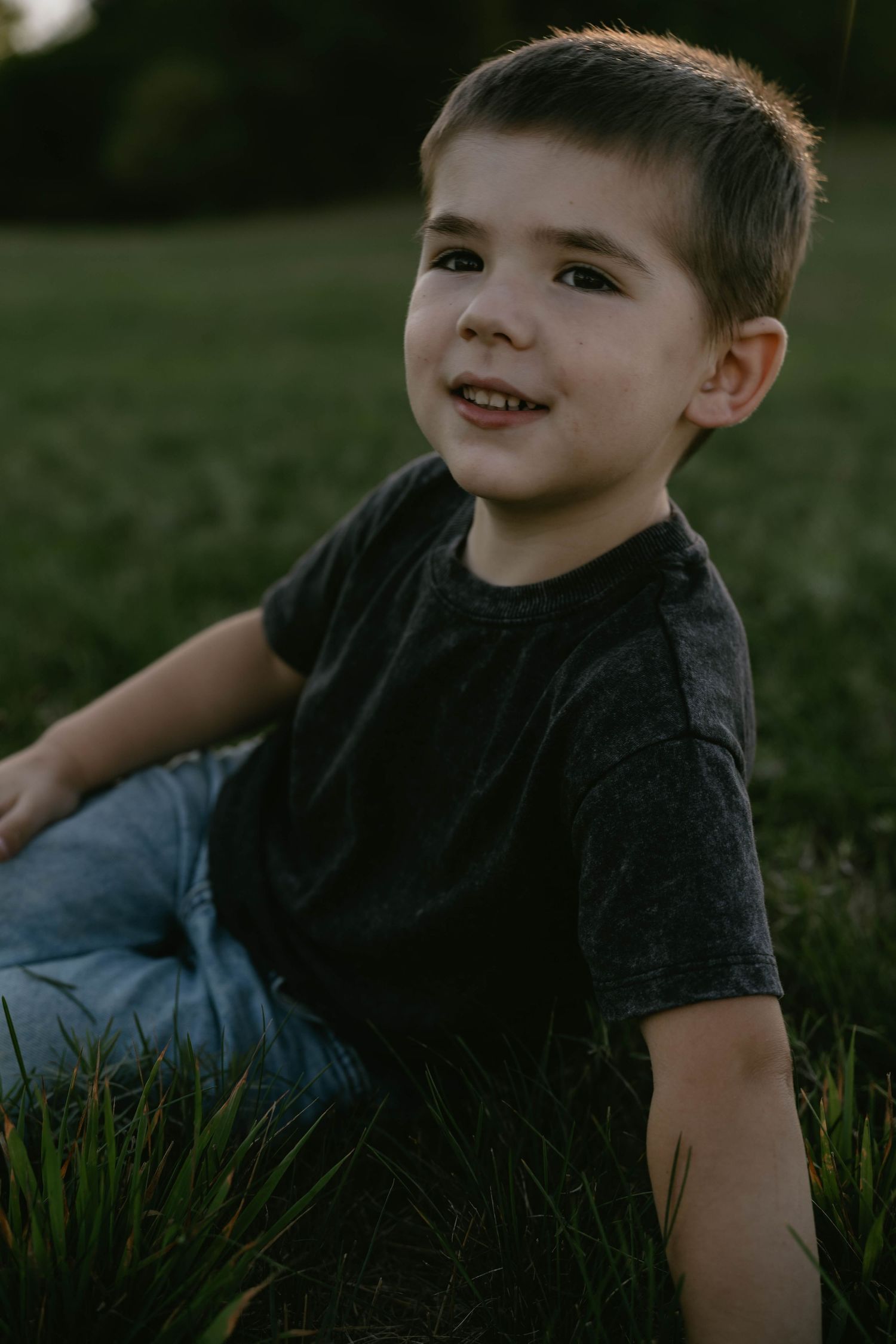 A child in a black t-shirt sits on grass during golden hour with a gentle smile.