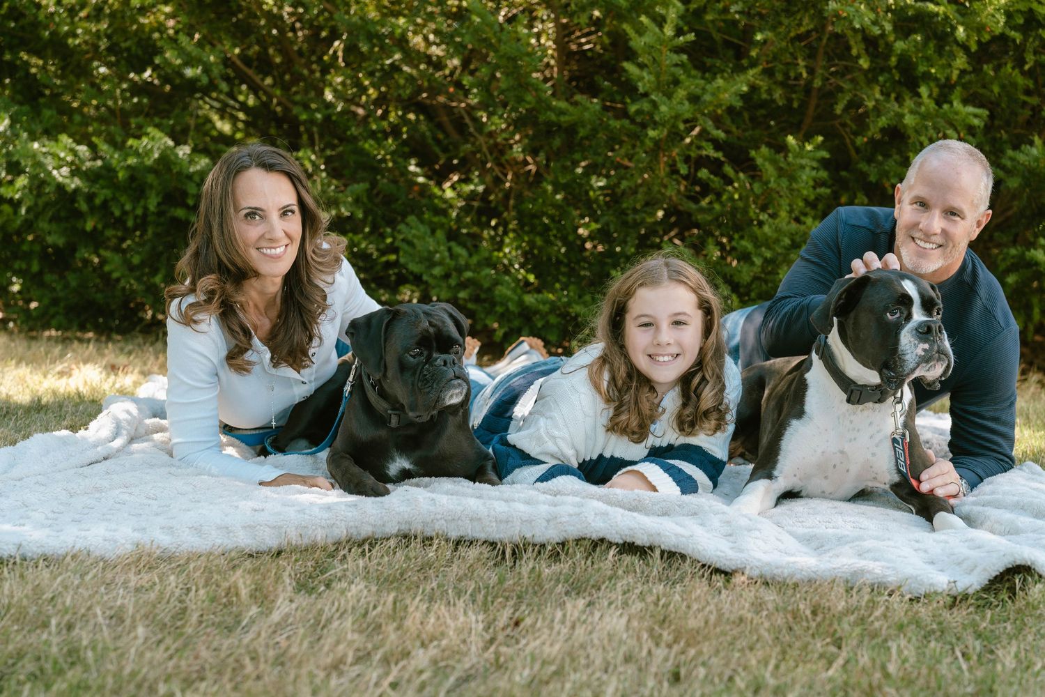 Family laying on white blanket during outdoor photo session with green foliage background.
