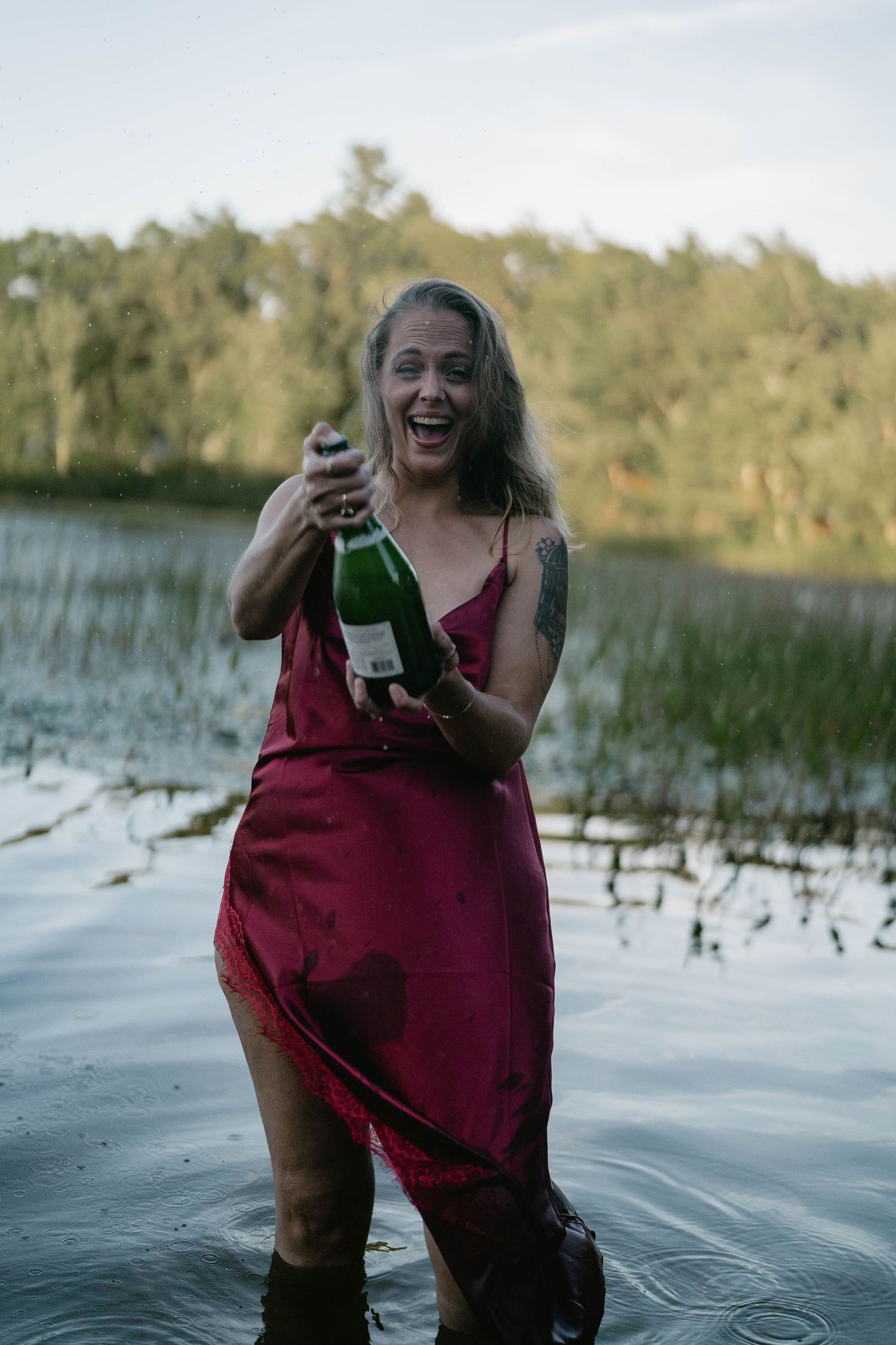 A person wearing a burgundy dress laughs joyfully while standing in a lake with marsh grass in the background.