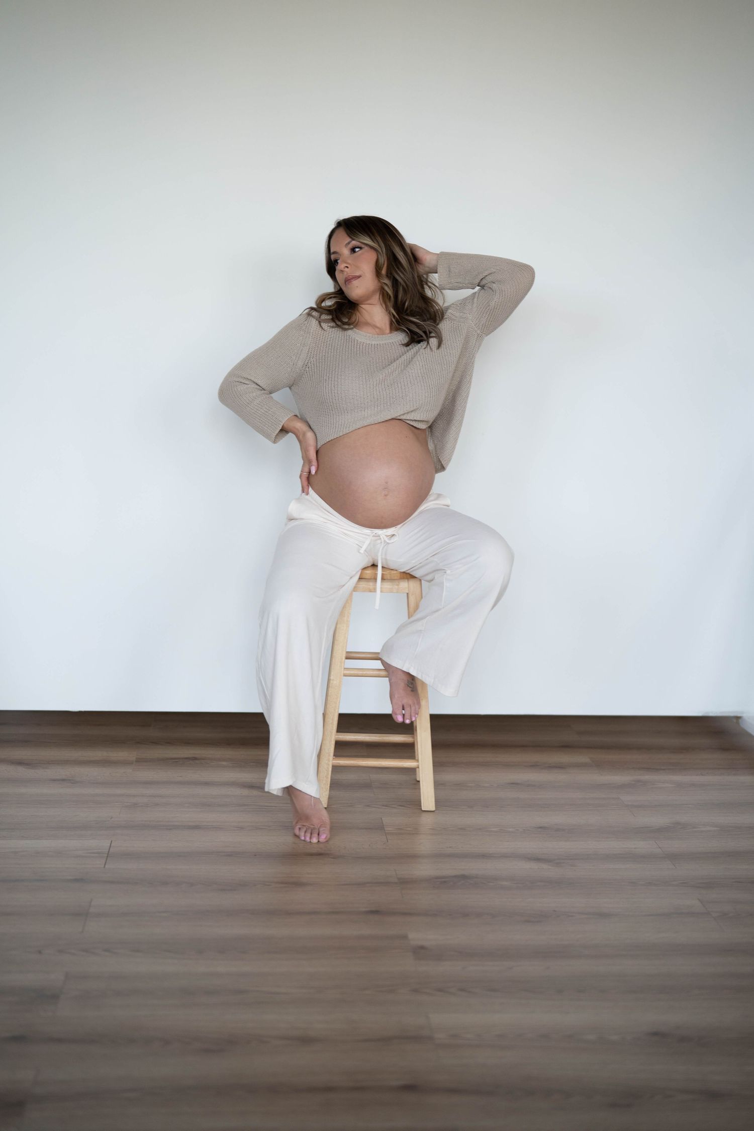 Expectant mother in beige sweater and white pants poses on wooden stool.