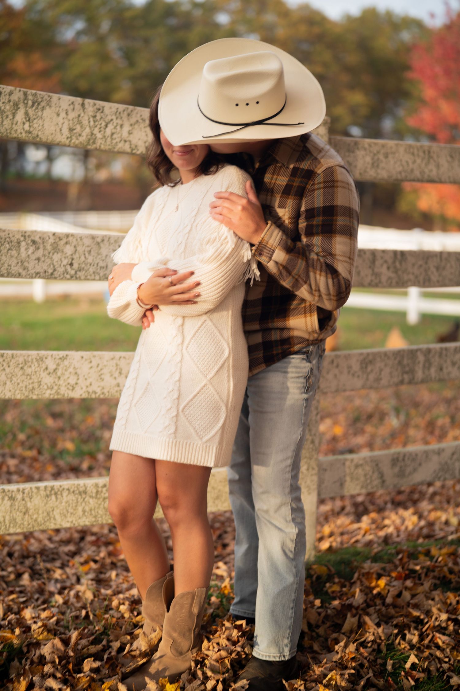 A romantic autumn scene with a couple embracing by a white fence surrounded by fallen leaves.