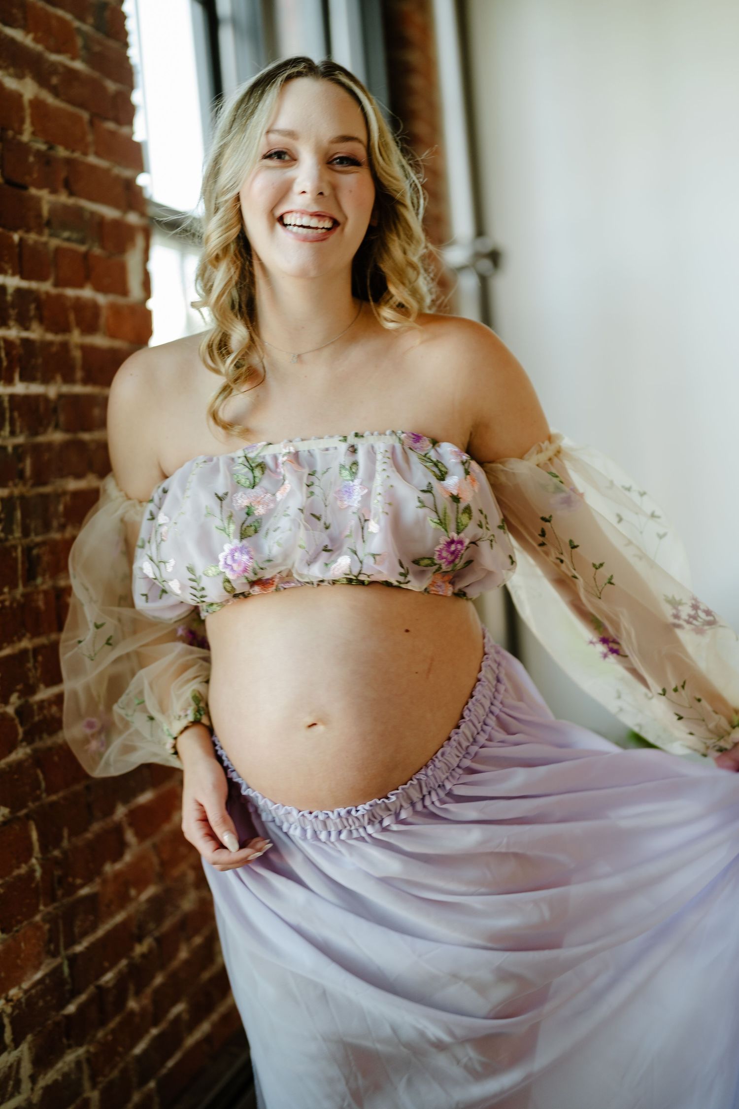A maternity photoshoot in front of a brick wall features a floral off-shoulder dress with delicate ruffled details.