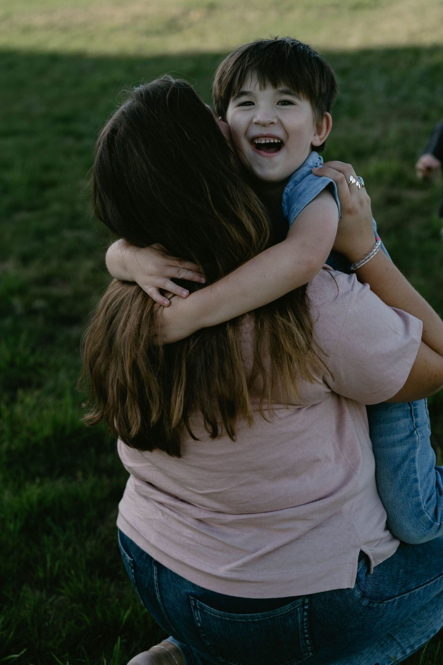 A joyful moment captured outdoors as a child in denim gets a loving embrace in a grassy field at sunset.