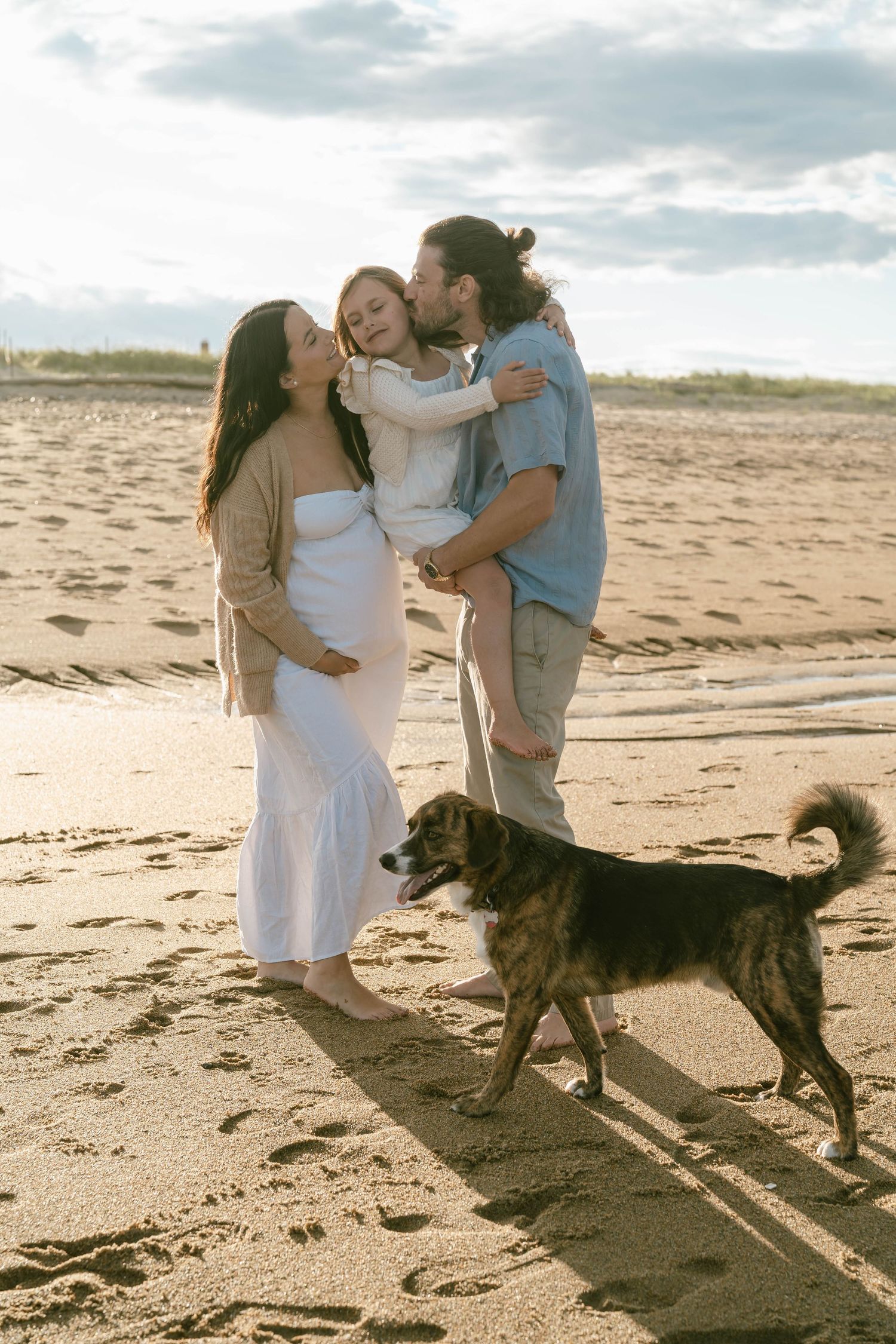A couple embraces on a sandy beach at sunset with their large black dog beside them.