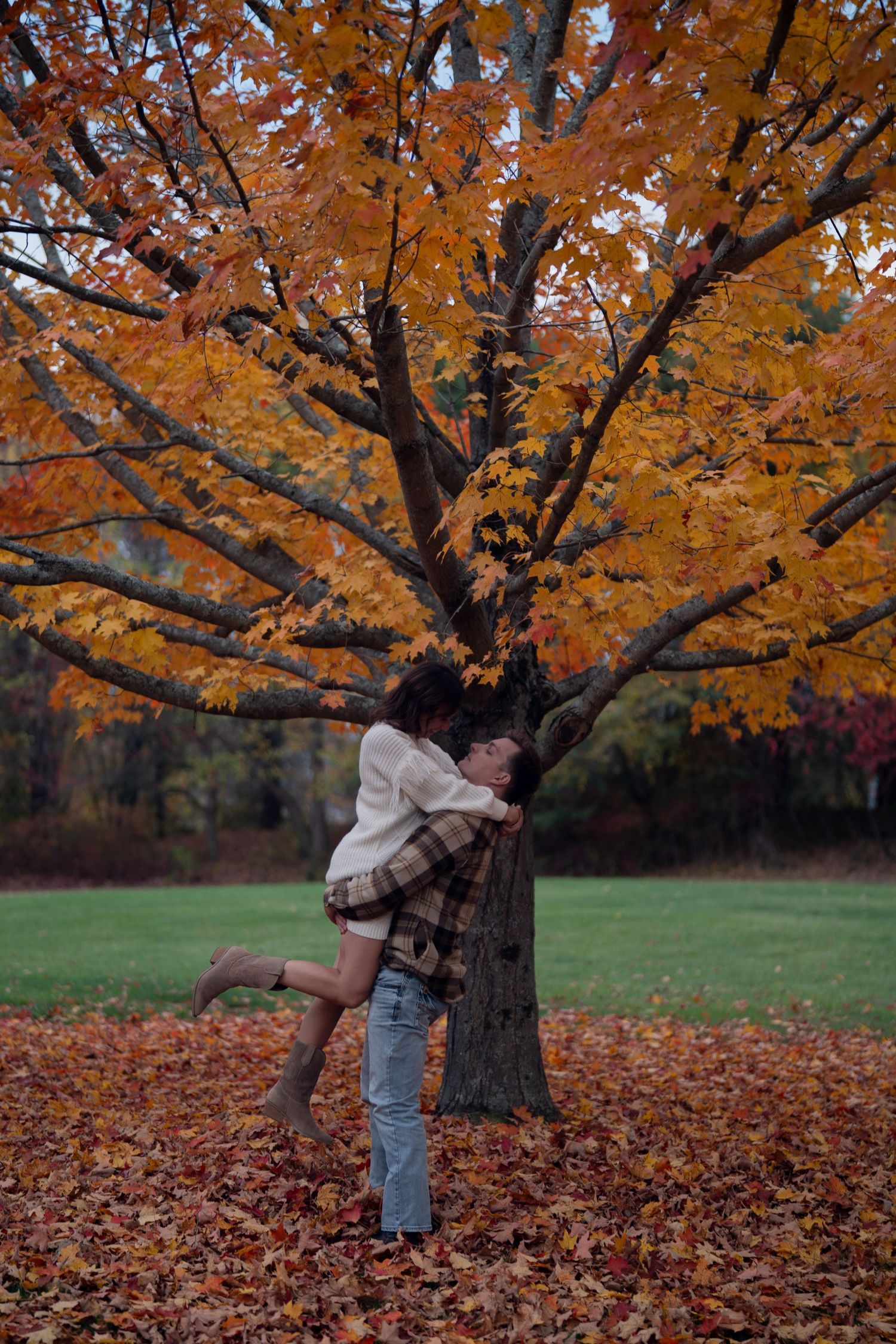 A tree with vibrant orange autumn foliage stands in a park with fallen leaves scattered beneath.