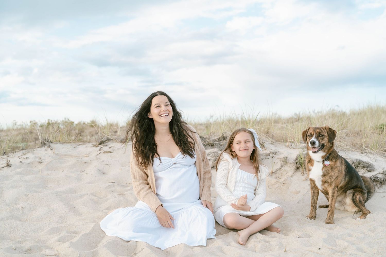 Beach photoshoot scene with people in white dresses and a dog on sandy dunes.