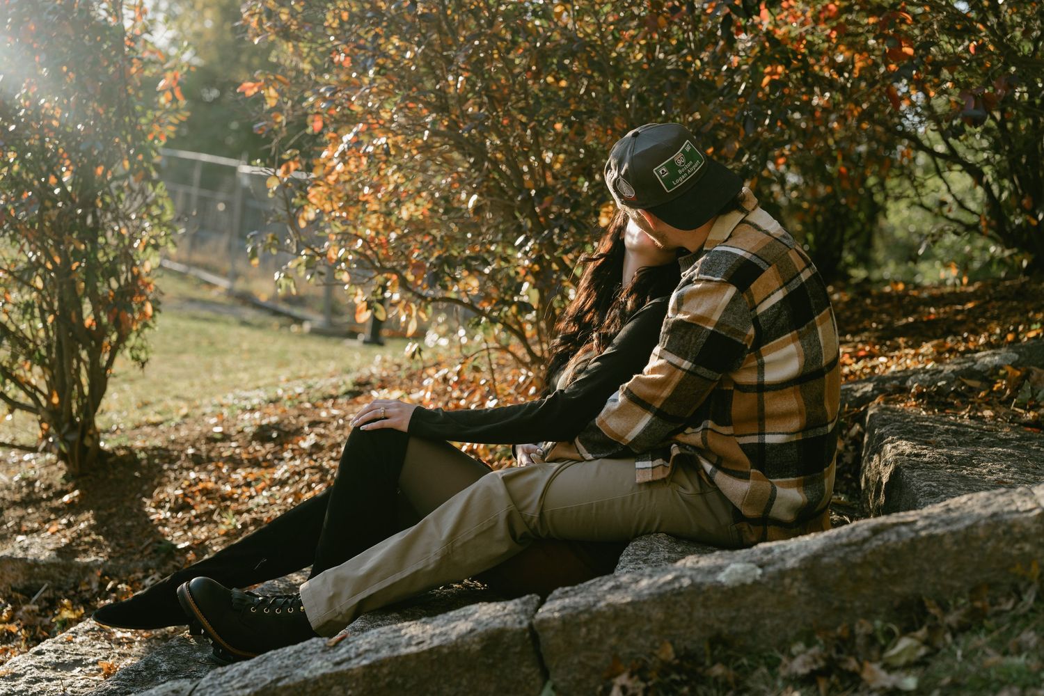 Two people share an intimate moment while sitting on fallen leaves in an autumn forest setting.
