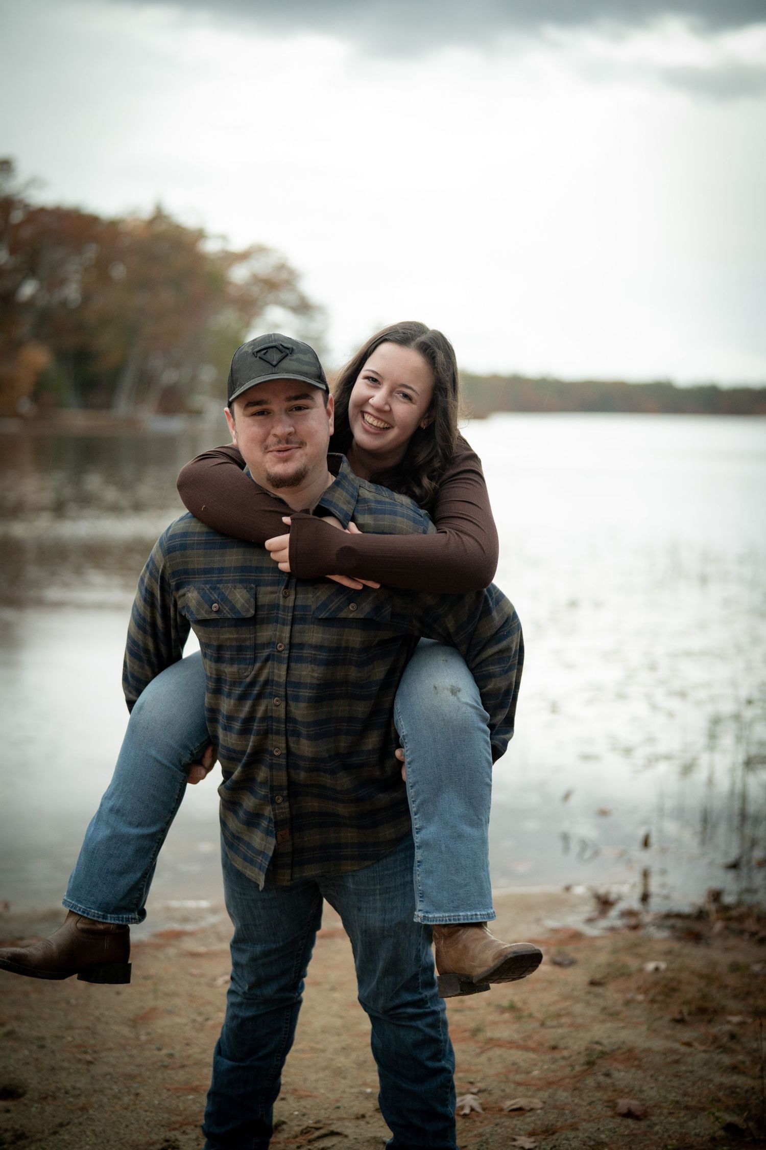 Playful couple enjoying a piggyback ride by the lake on cloudy day.