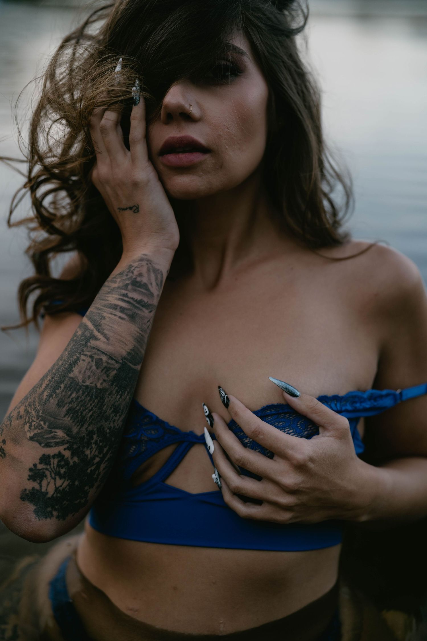 Artistic portrait showcasing intricate arm tattoos and blue attire against a waterfront backdrop.