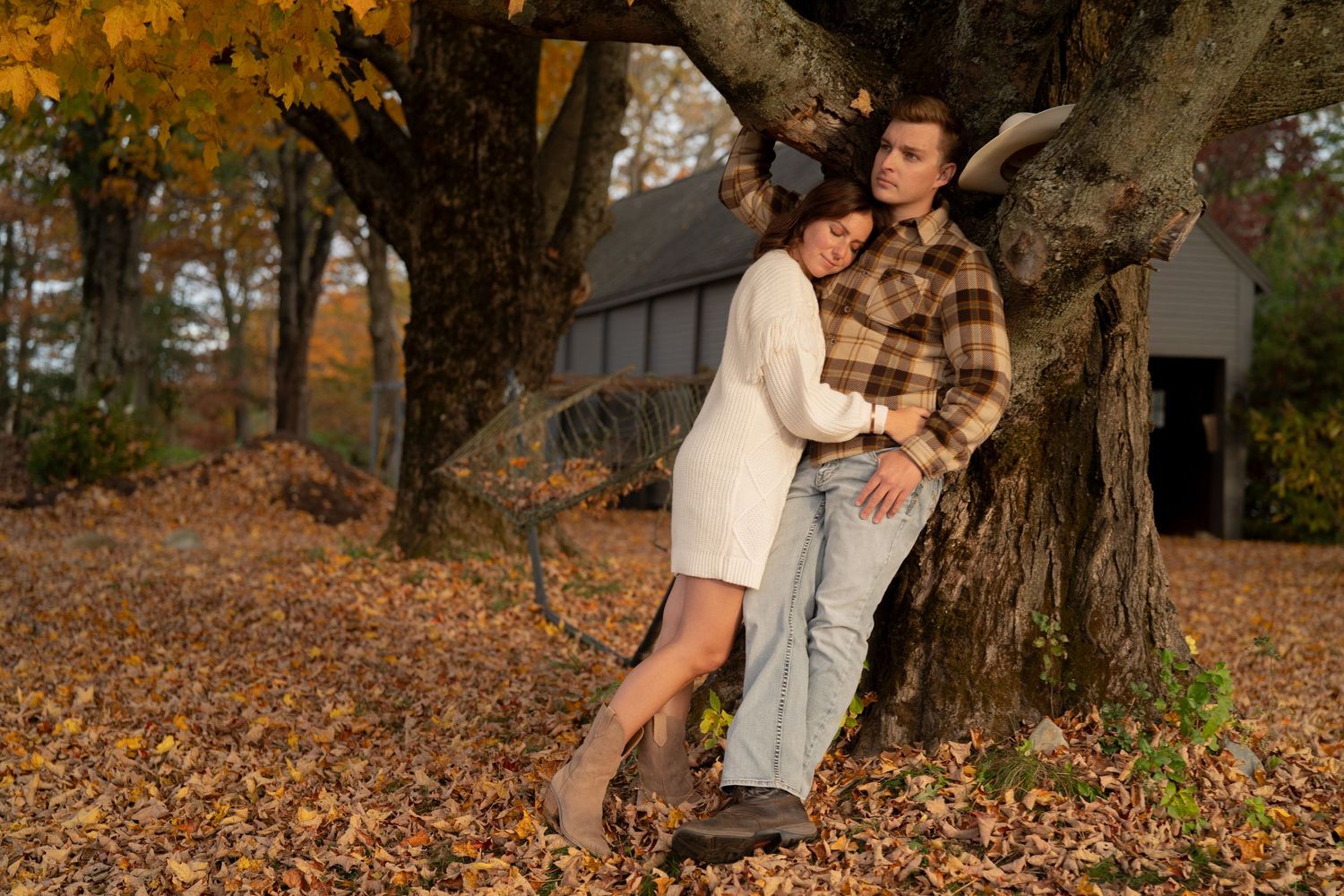 Couple in casual attire poses together among autumn trees and fallen leaves in a romantic outdoor photoshoot.