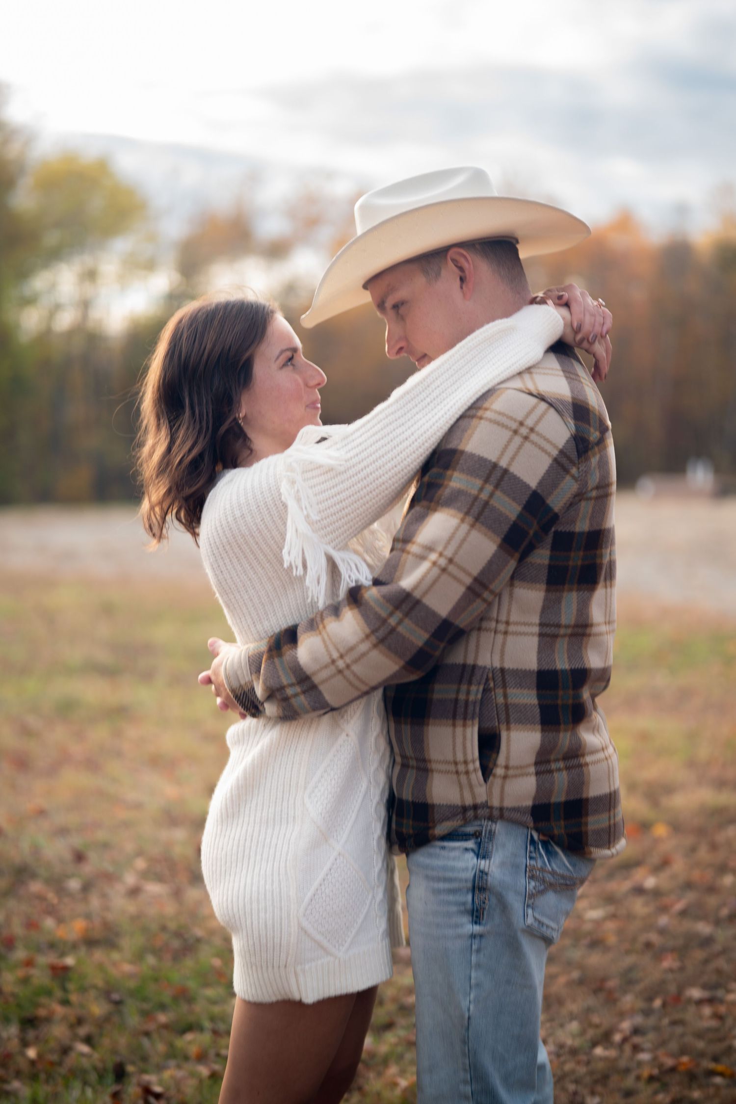 A couple embraces while standing in a field during golden hour wearing casual fall attire.