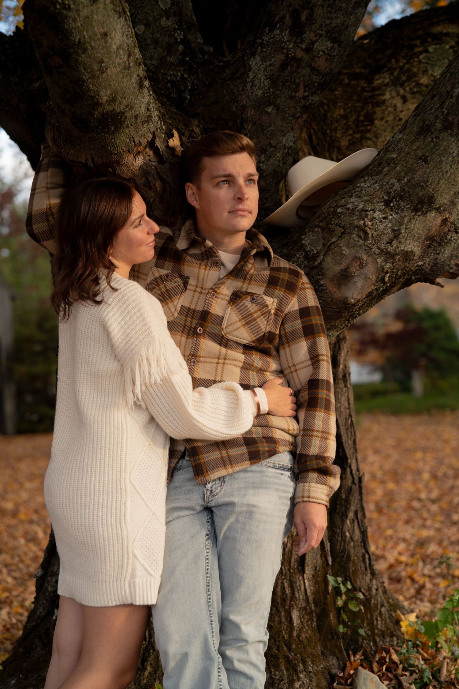 A couple in autumn attire embraces against a tree trunk surrounded by fallen leaves.