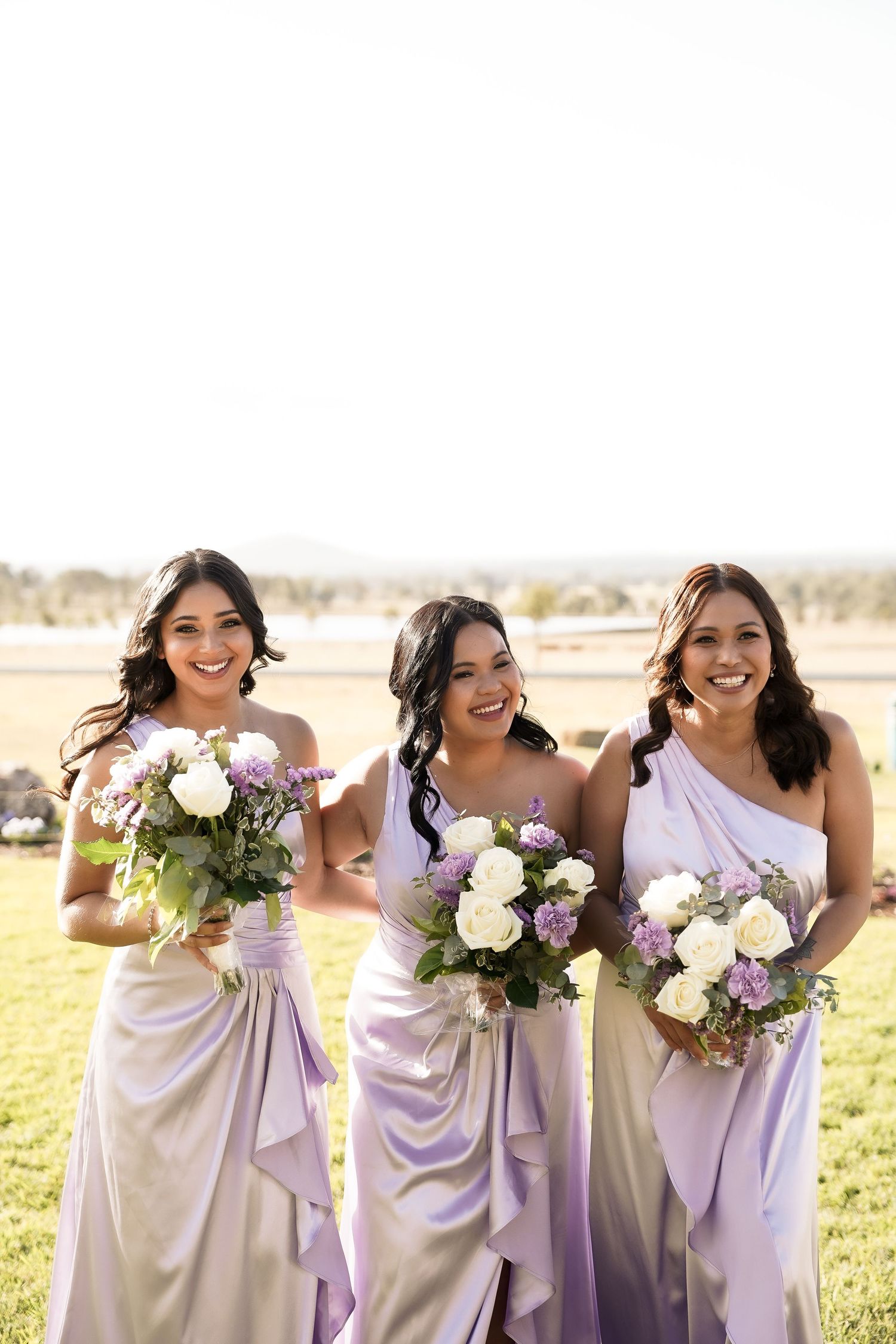 Bridesmaids in lavender dresses pose with white floral bouquets in golden sunlight.
