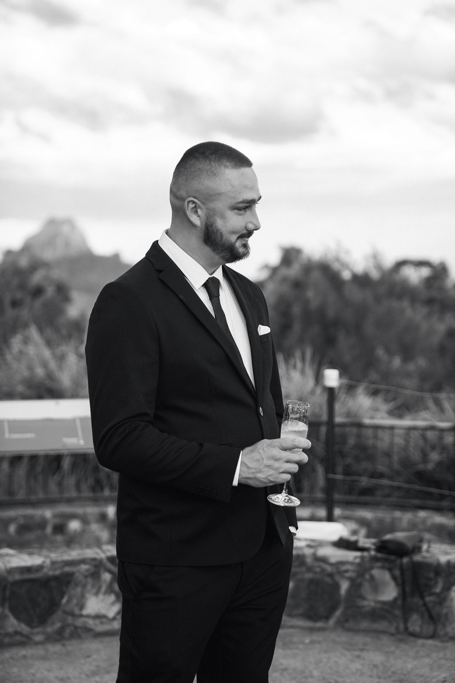 A person in a black suit holds a wine glass while standing outdoors against a scenic mountain backdrop.