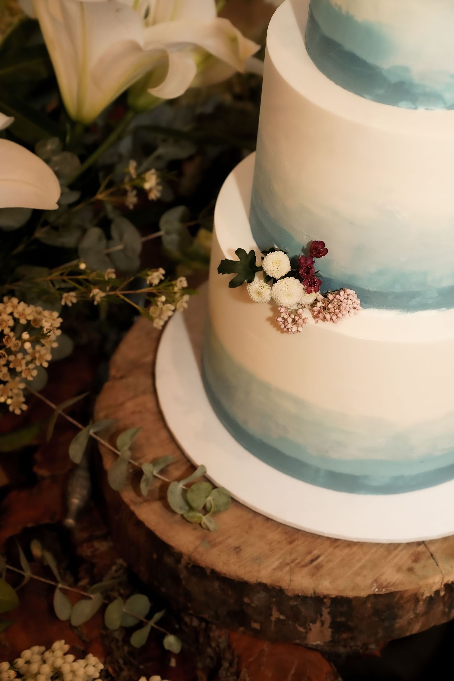 An elegant white wedding cake with blue watercolor details displayed on a rustic wood slice.