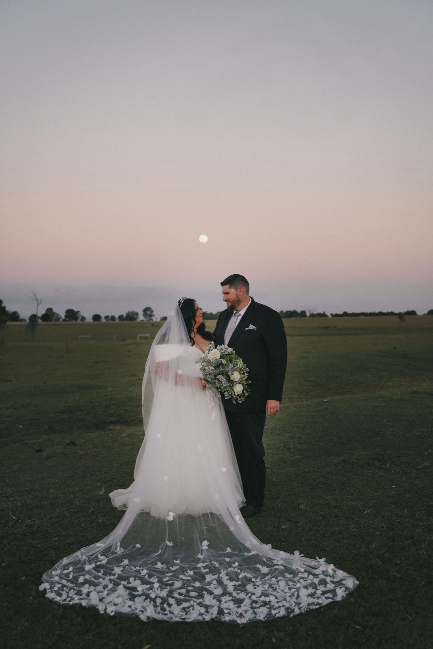 A bride and groom share an intimate moment at sunset in an open field with her flowing white dress spread on the ground.