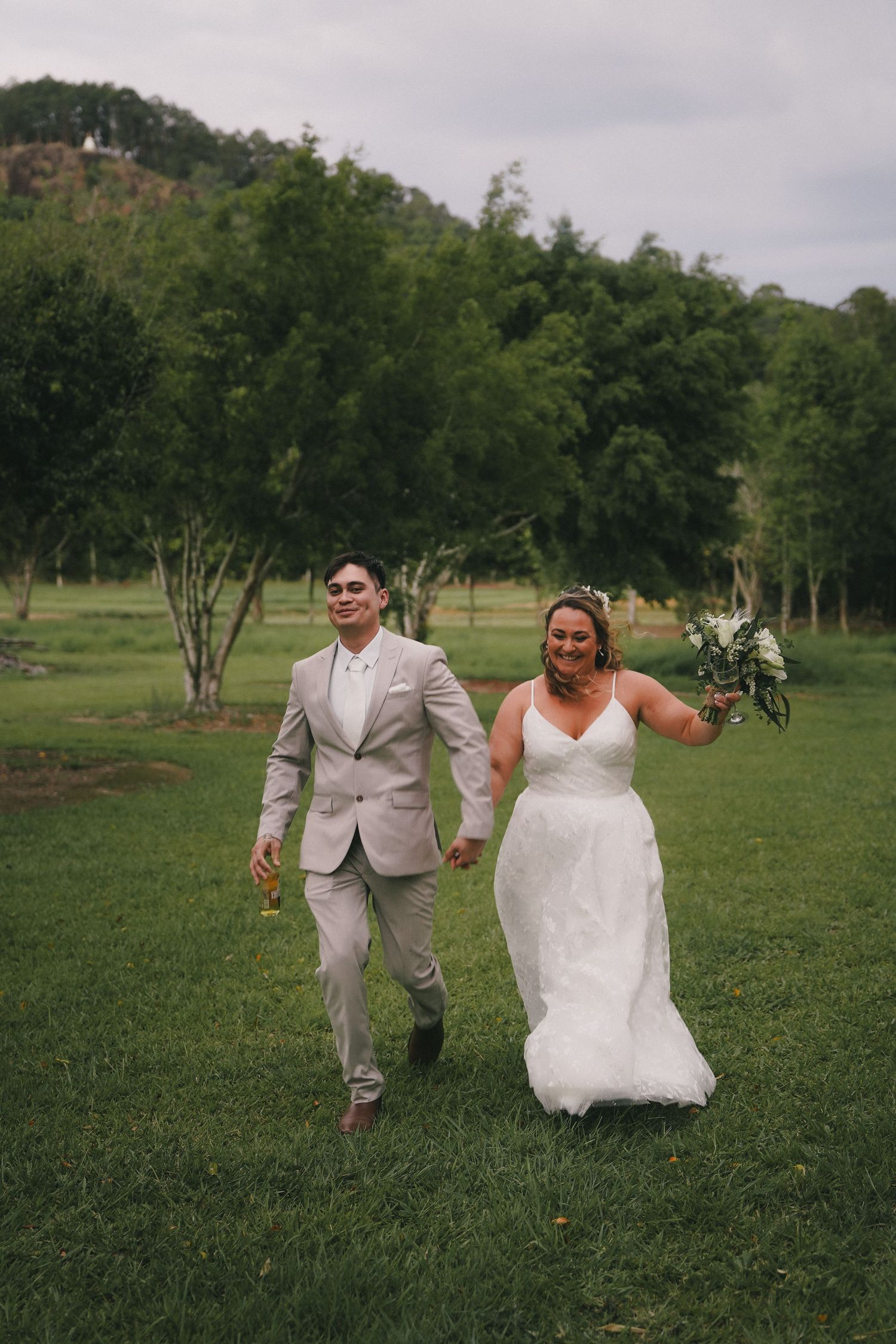 A couple in wedding attire runs joyfully across a green lawn with trees and hills in the background.