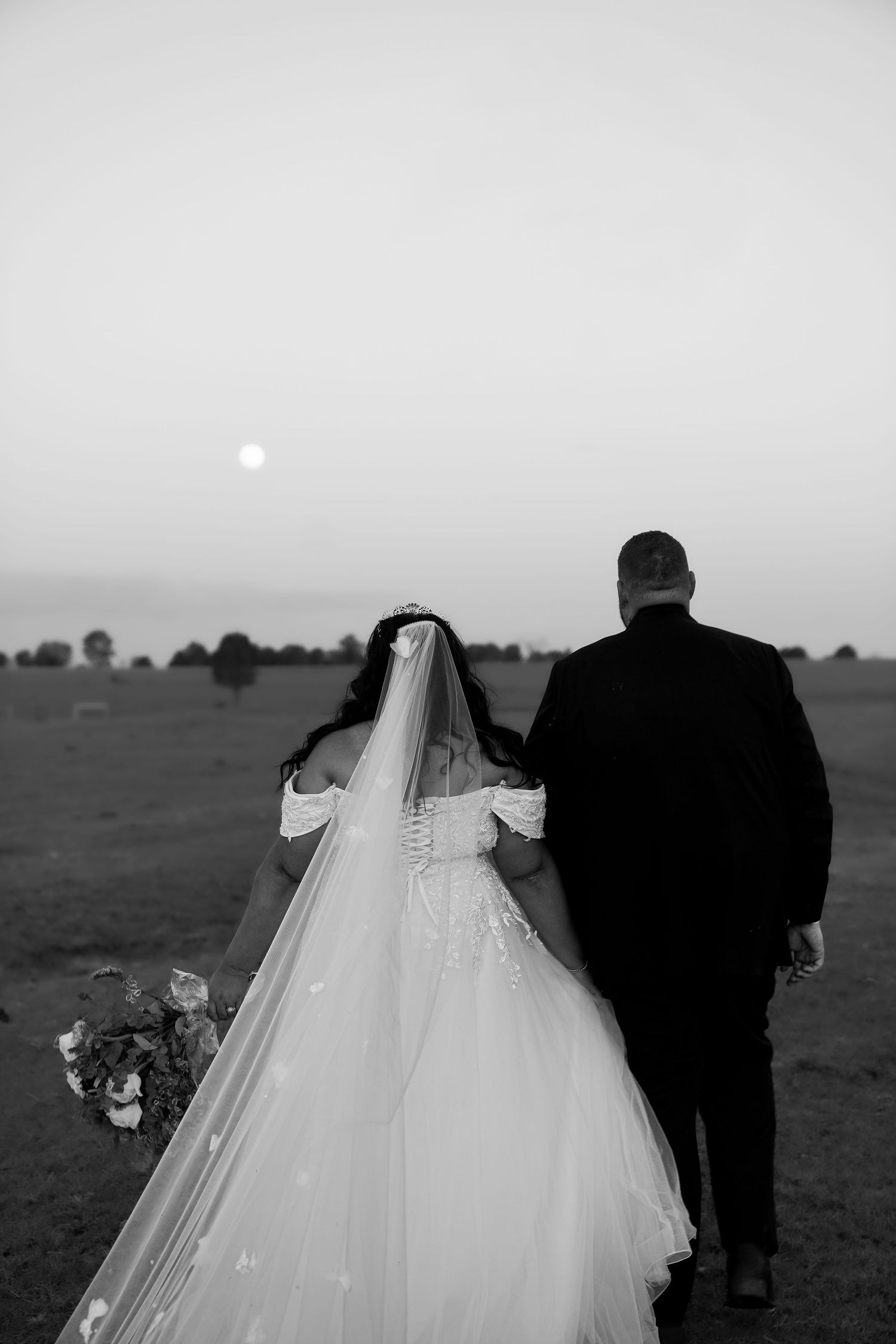 Black and white photo of a bride and groom walking in a field at sunset with a full moon visible in the sky.