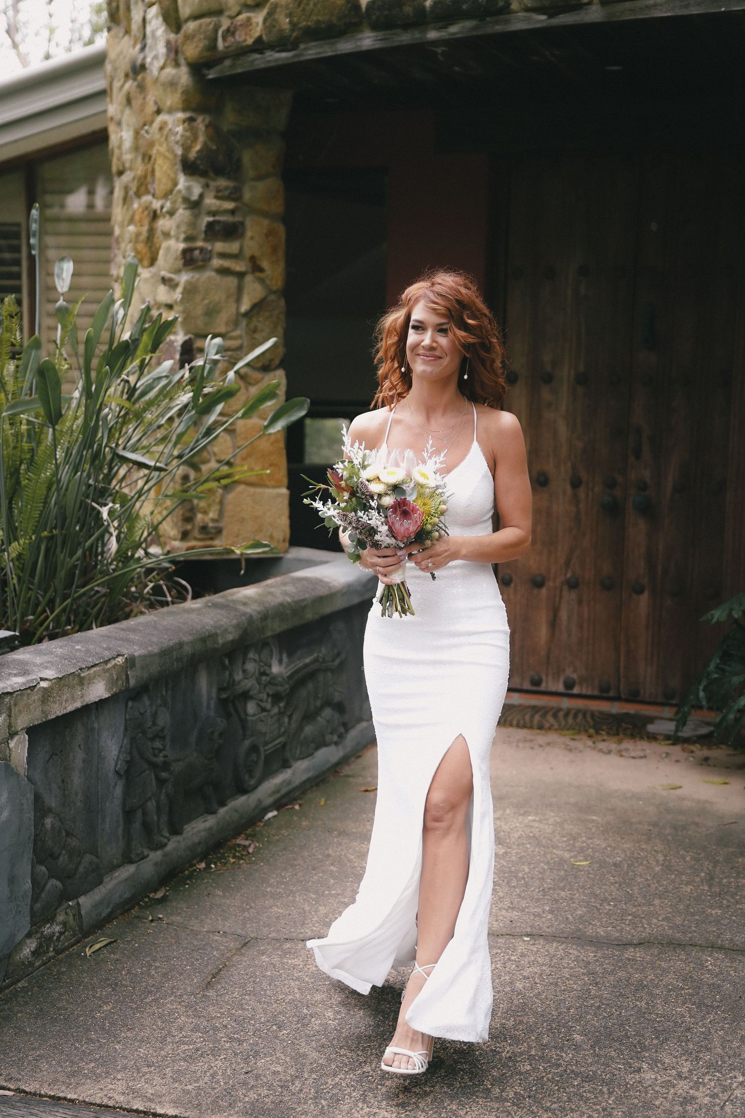 A bride in a white wedding dress poses on a stone walkway with a rustic wooden door and stone walls in the background.