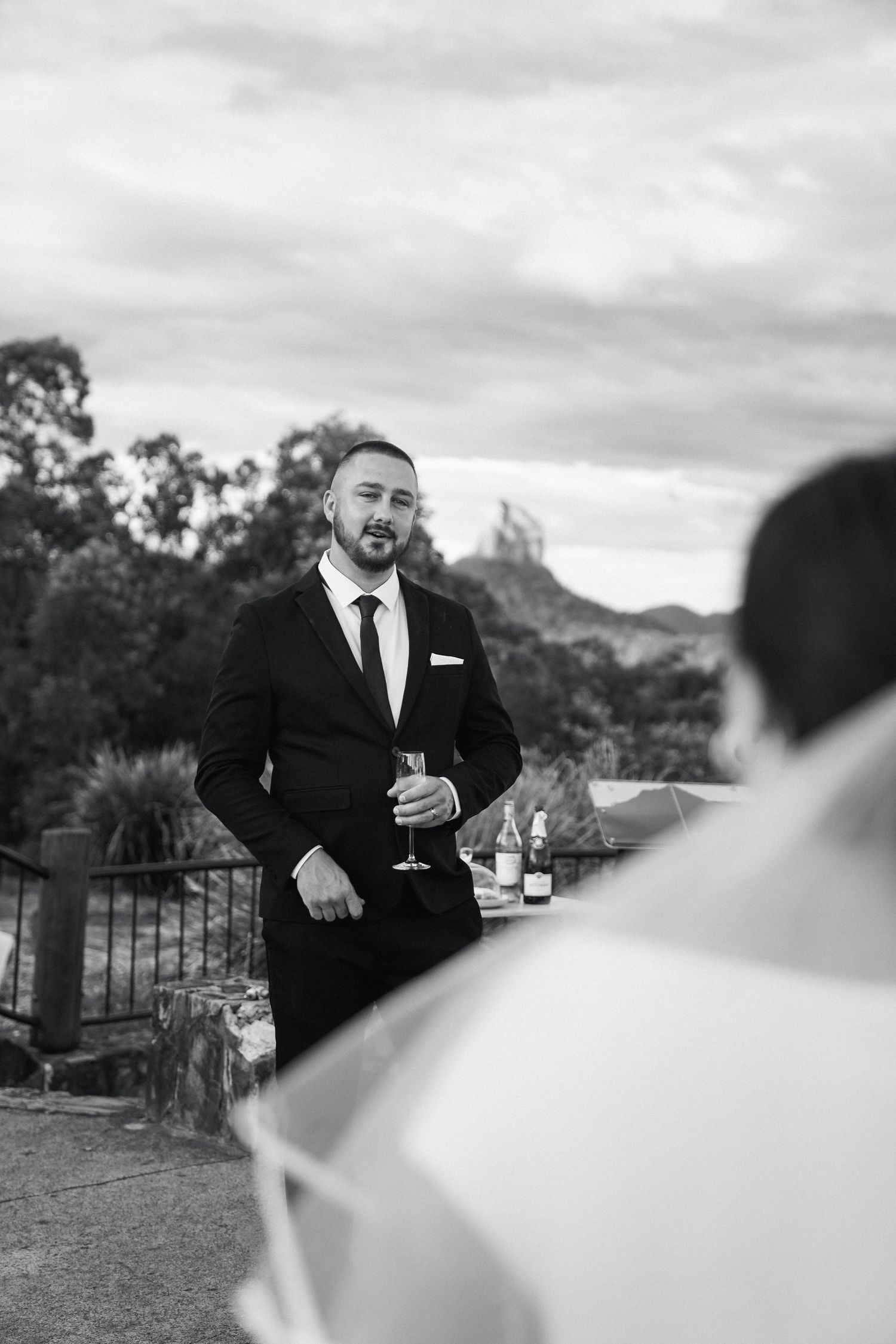 A person in a black suit holds a wine glass on an outdoor terrace with mountains and trees in the background.