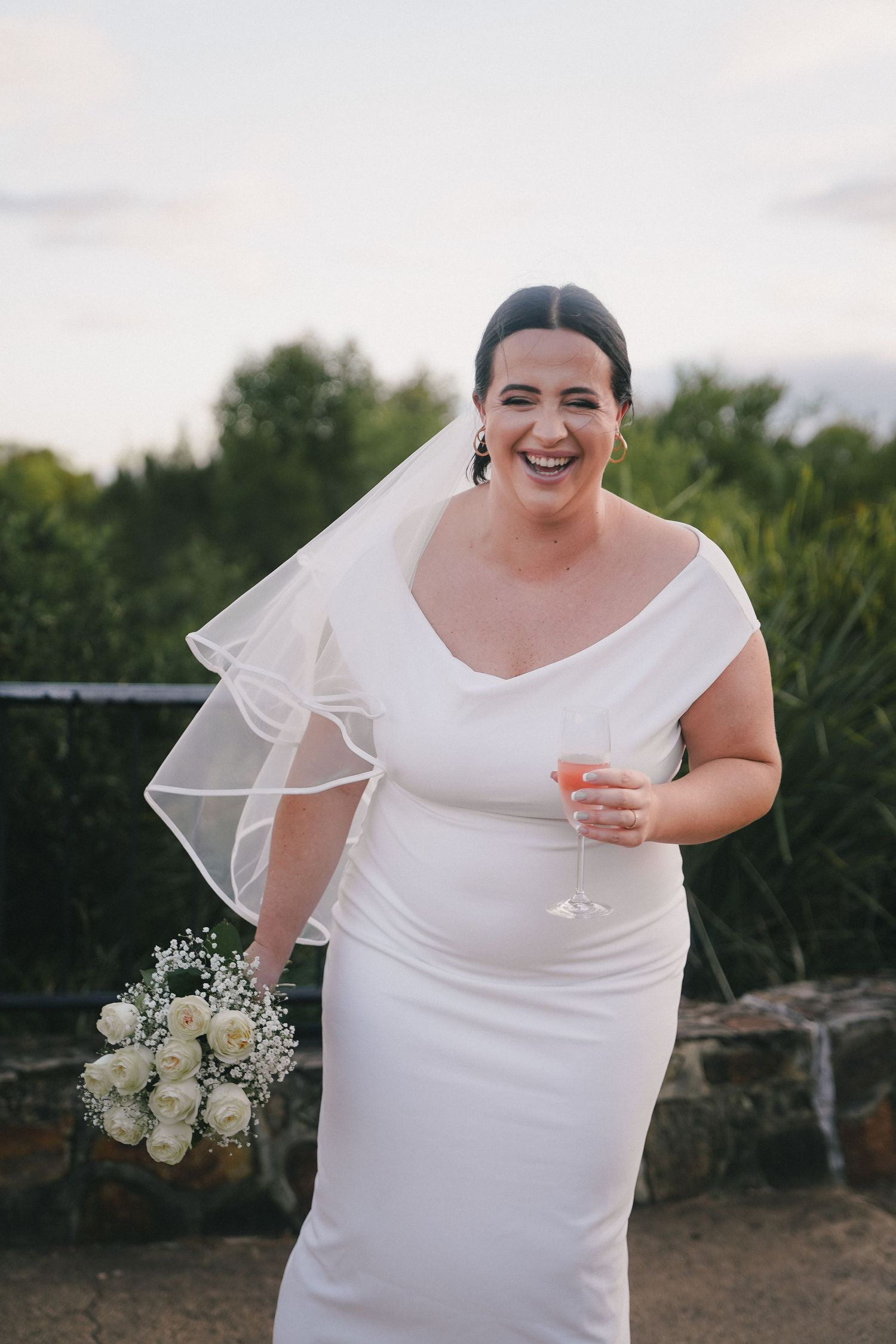 A joyful bride in a simple white wedding dress laughs while holding a white floral bouquet outdoors.