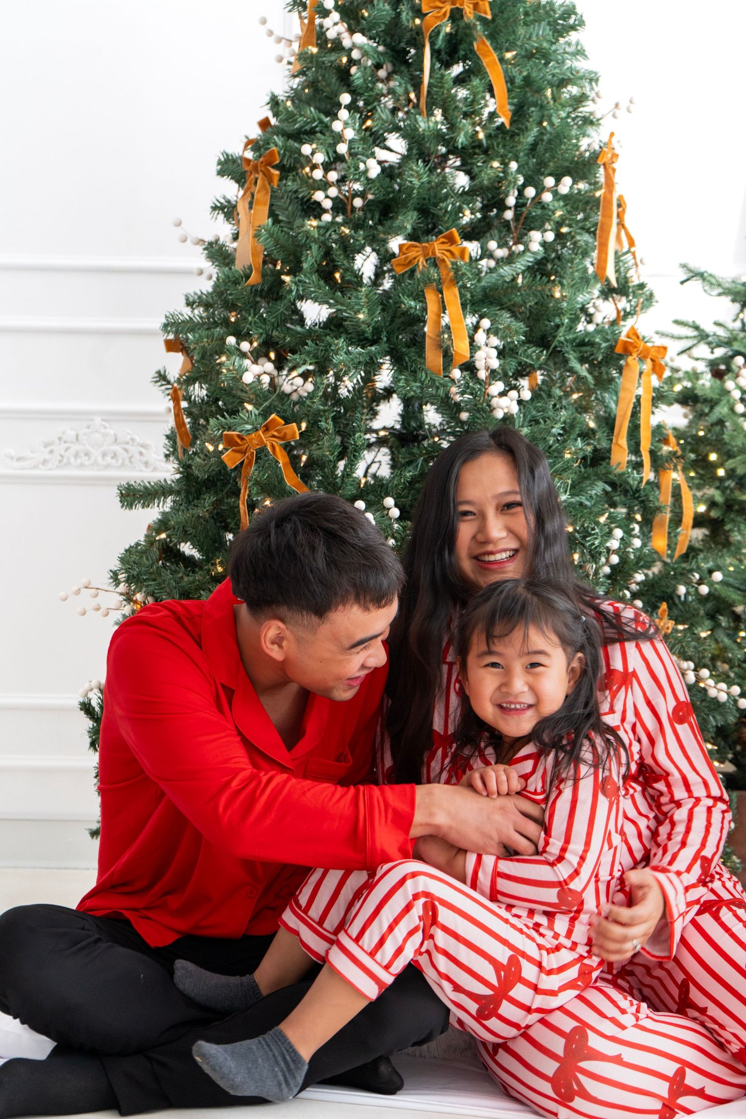 Asian family in red pajamas smiling together in front of decorated Christmas tree with gold ribbons.
