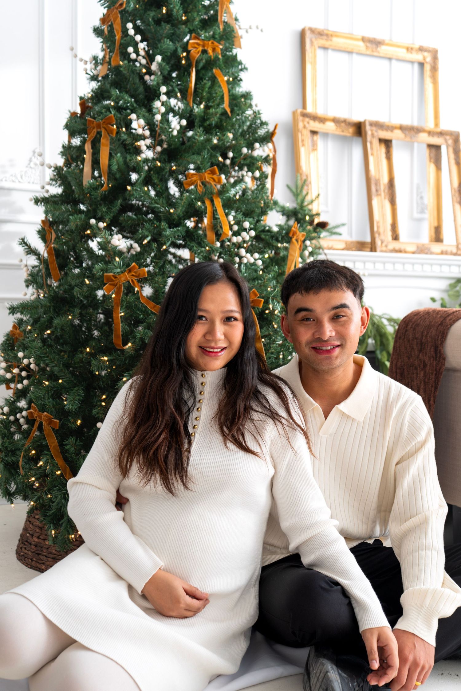 Smiling couple in white outfits sitting together in front of decorated Christmas tree with gold ribbons.