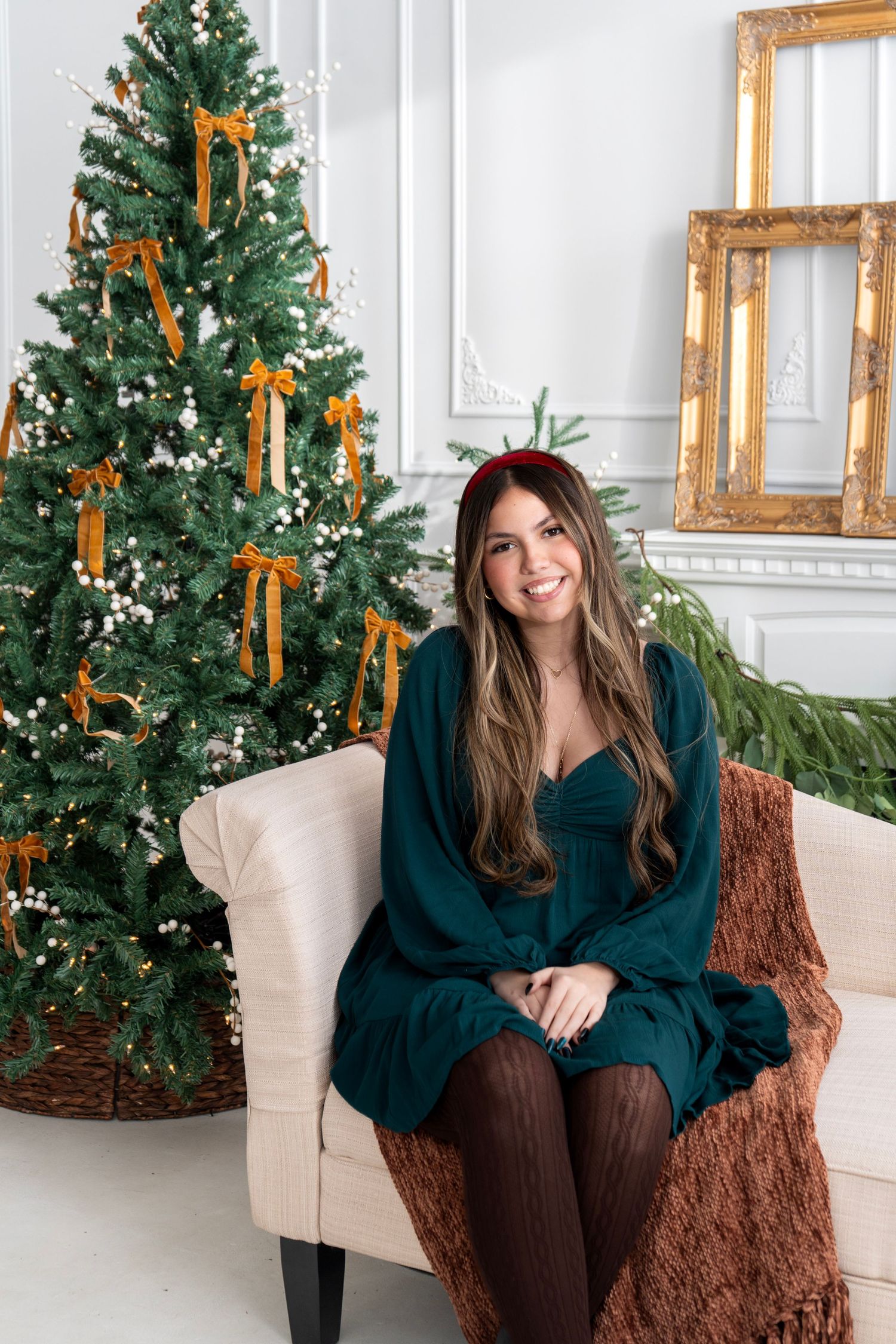 Woman in green dress and red headband smiles on cream sofa beside decorated Christmas tree with gold ribbons.