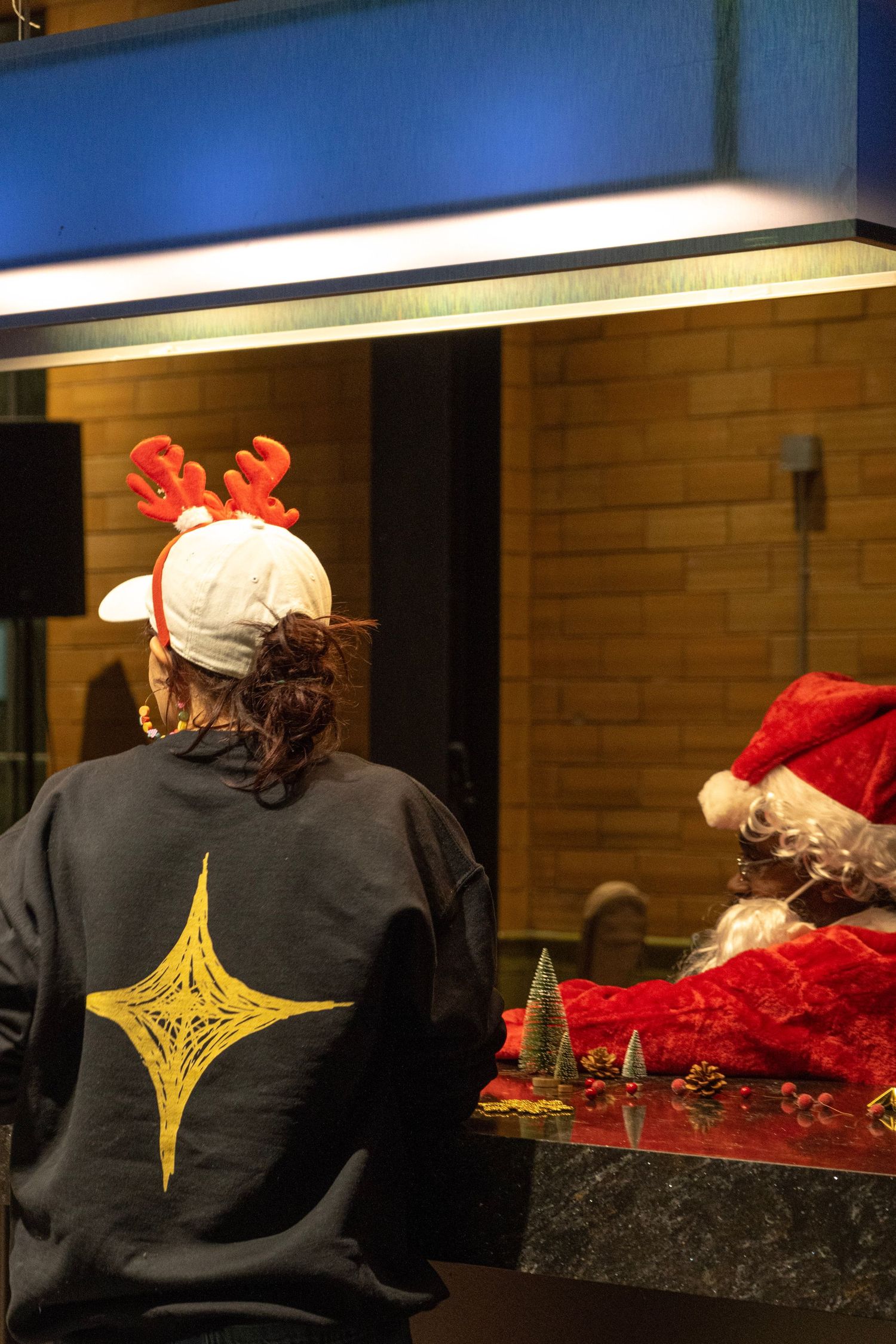 Person wearing reindeer antlers headband and star sweatshirt meets Santa Claus through a window at Christmas event.