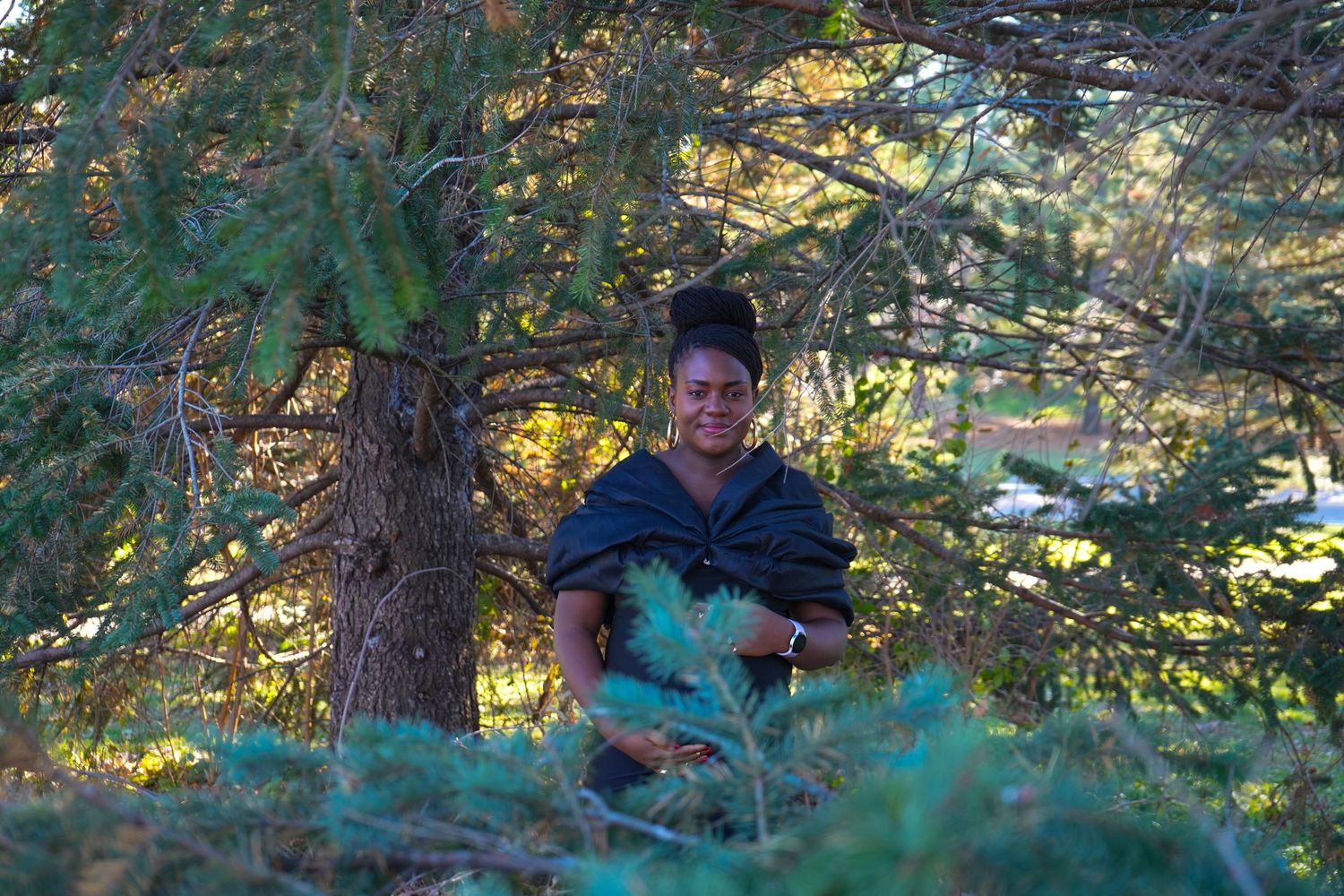 Woman standing among pine trees in a sunlit forest, holding green branches while wearing a dark wrap.