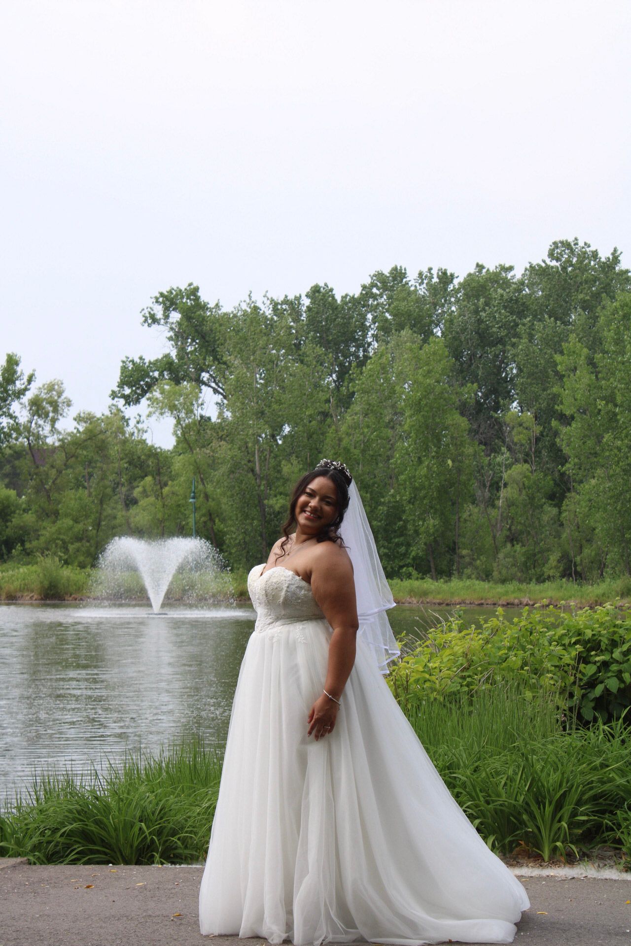 Smiling bride in white strapless gown and veil poses by a serene pond with fountain and lush green trees.