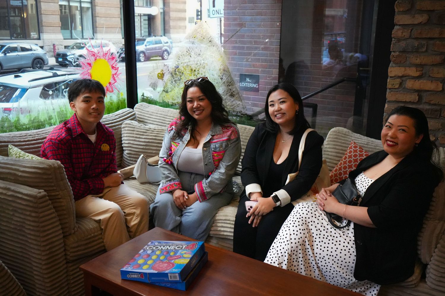 Four women smiling and sitting together on a couch in a cozy lounge setting with a board game on the table.