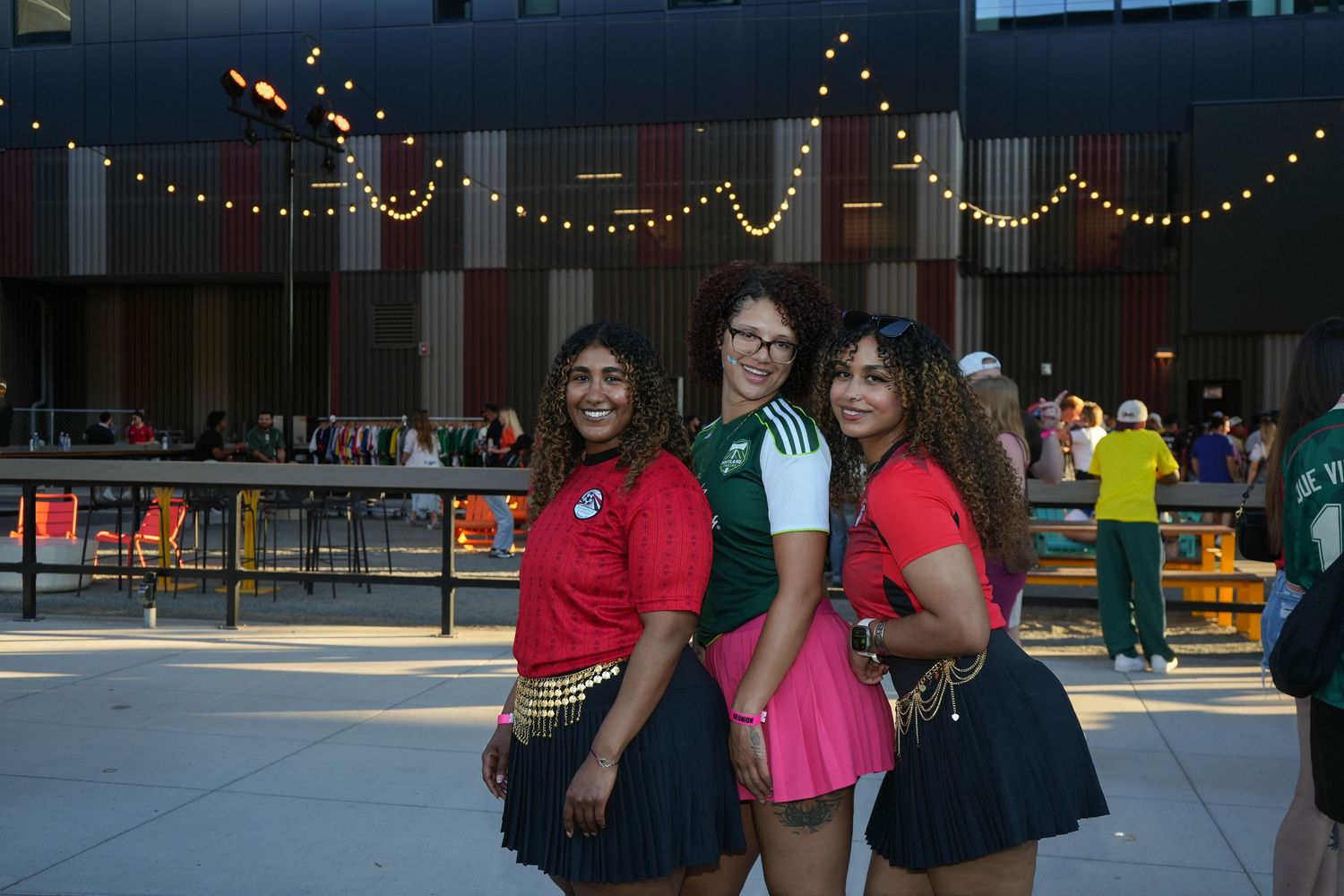 Three young women posing together outdoors at a festive evening event with string lights in the background.