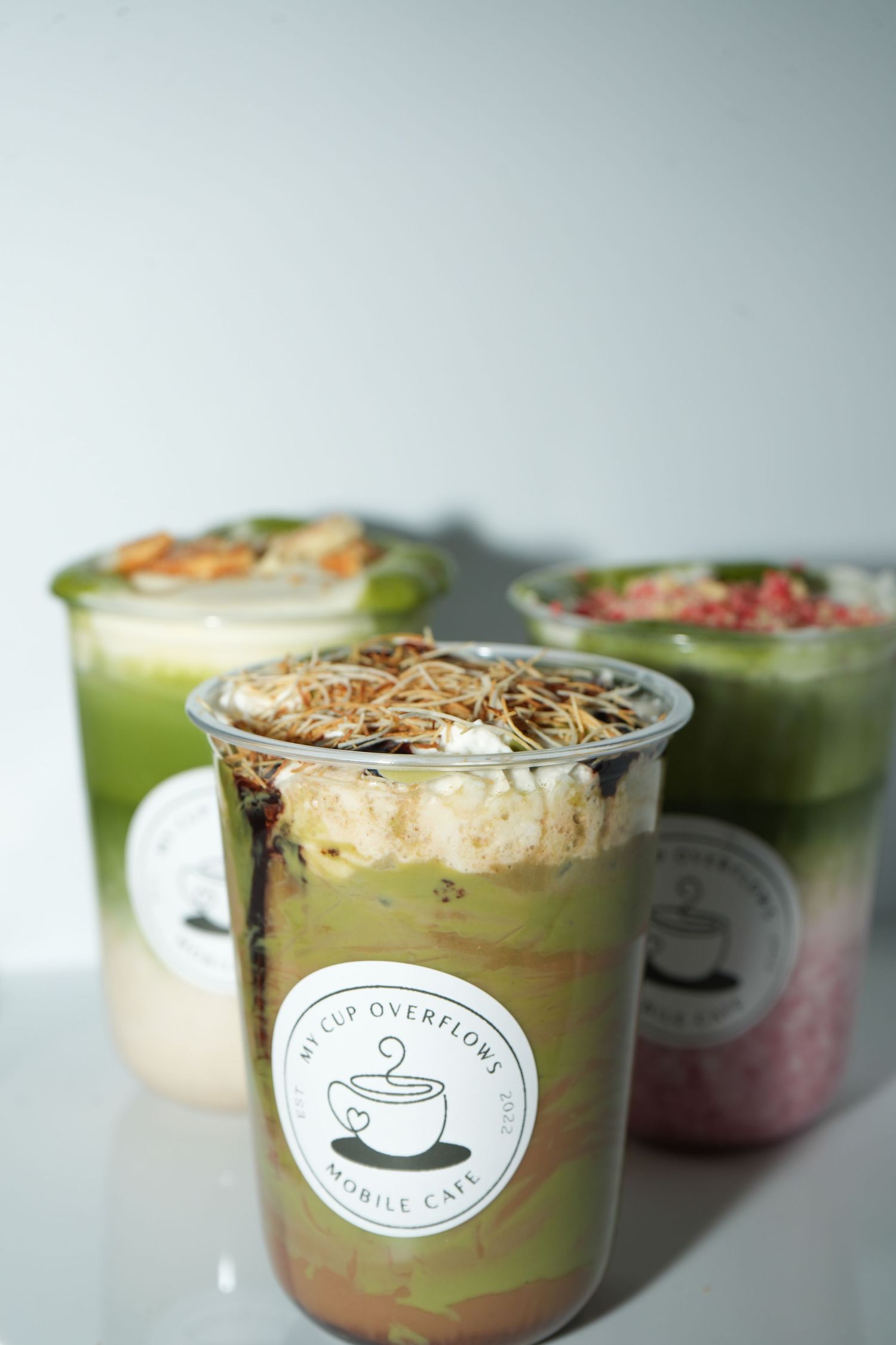 Three layered matcha drinks in clear cups with toppings, branded My Cup Overflows Mobile Cafe.