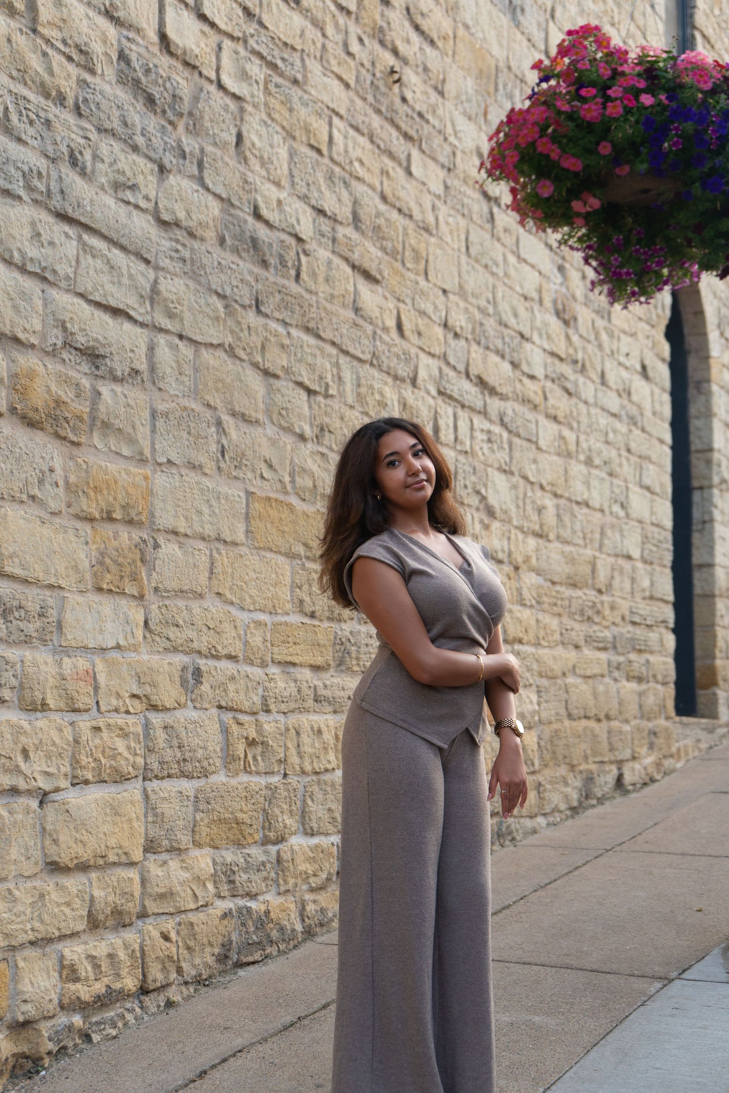 A young woman in a taupe jumpsuit poses gracefully beside a stone wall with colorful hanging flowers.