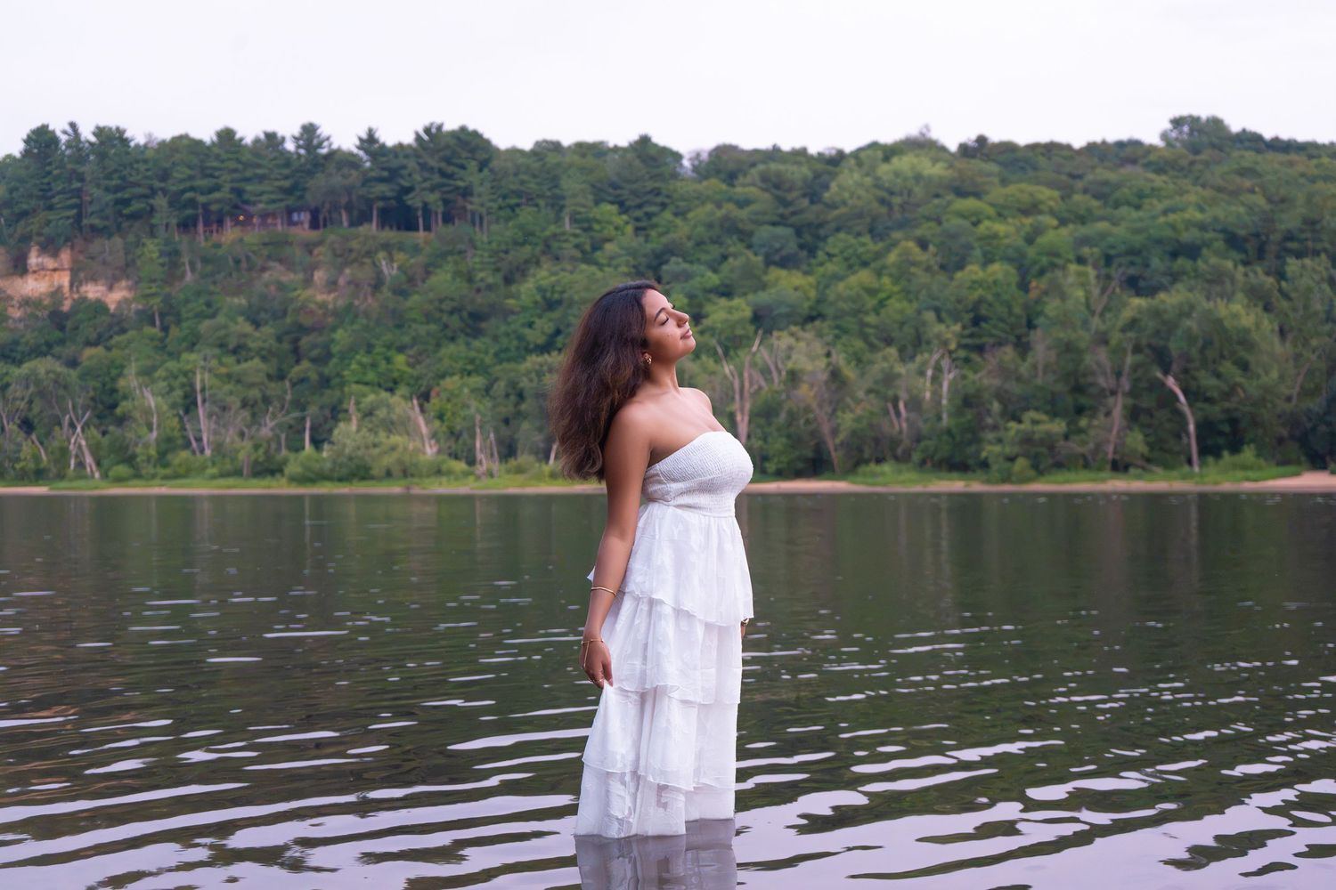 Woman in white dress standing in a calm river with lush green forested hills in the background.