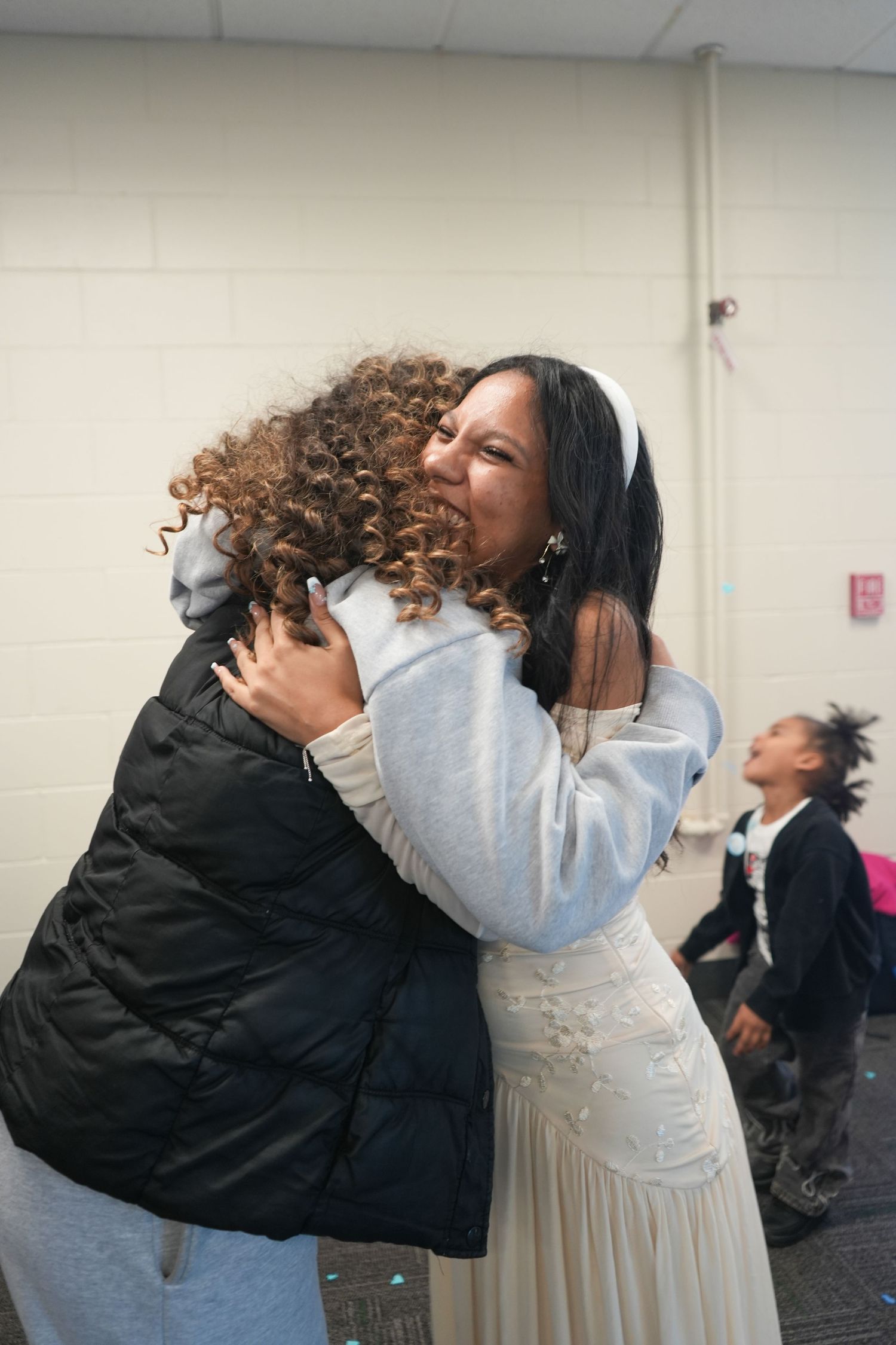 Two women sharing an emotional hug in a community center, one wearing a white dress and headband.
