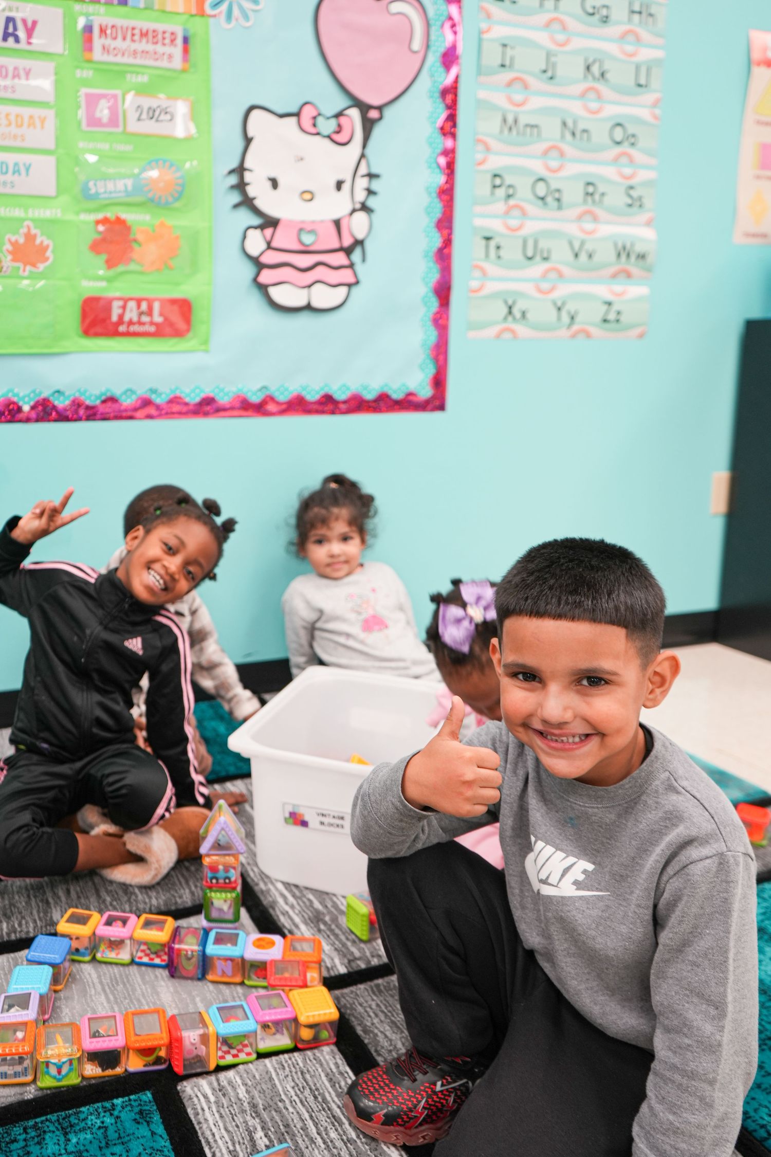 Children smile and give thumbs up in a colorful classroom with Hello Kitty decor and alphabet charts on the wall.