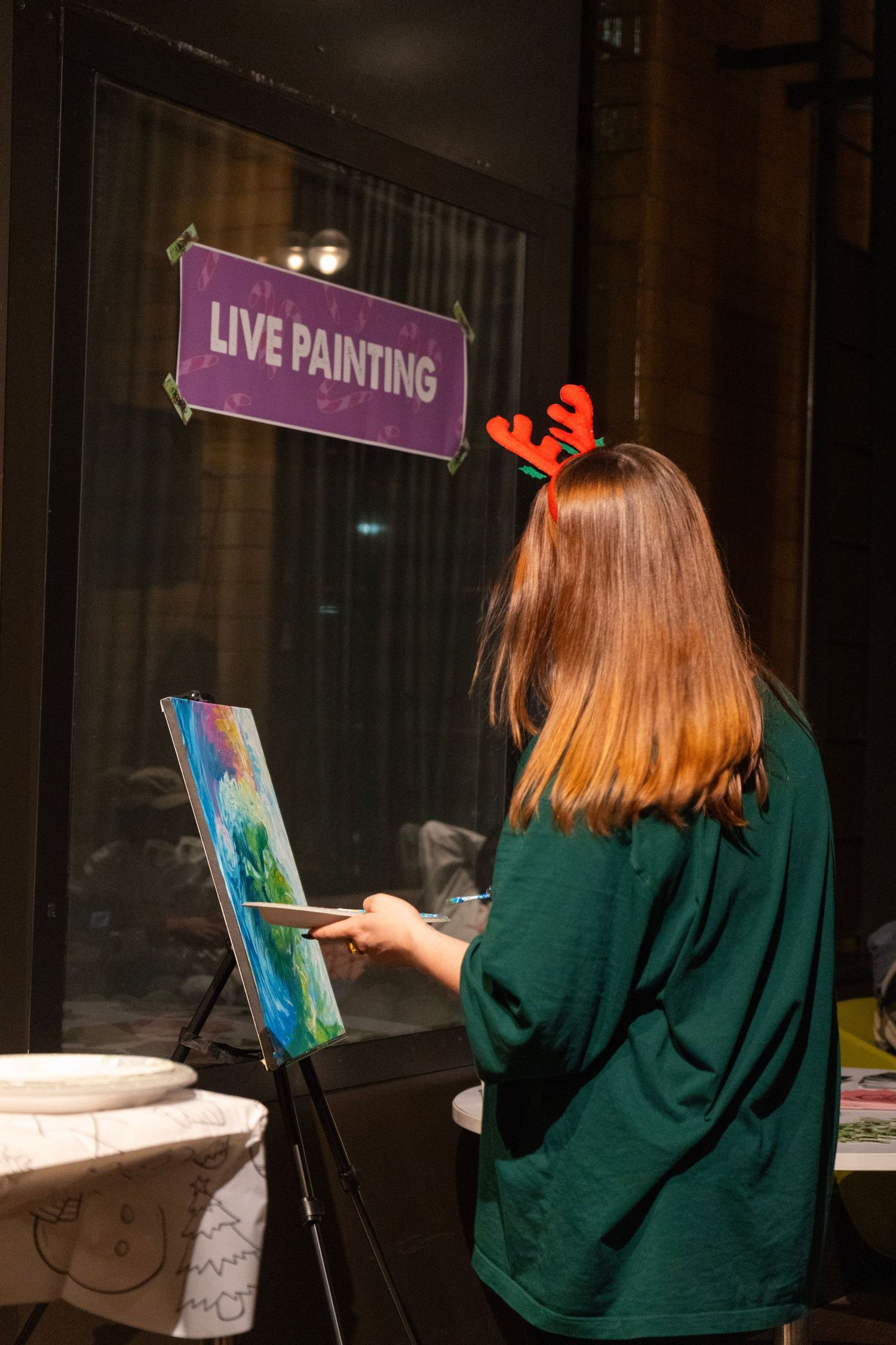 Woman with reindeer antlers painting on easel at a live painting event in a dimly lit venue.